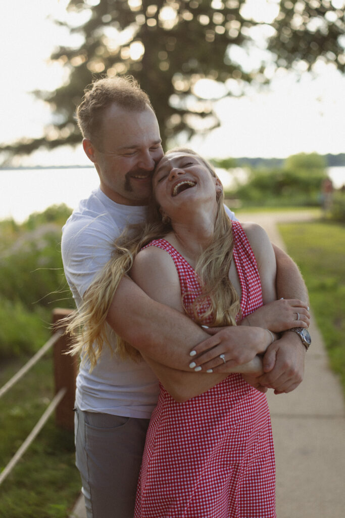 Guy holding his girl from behind whispering sweet nothings in her ear as she laughs throwing her head back during their engagement photos in Excelsior