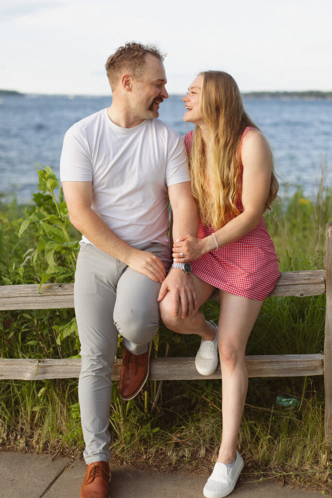 Engagement couple sitting on a wooden fence in front of lake Minnetonka