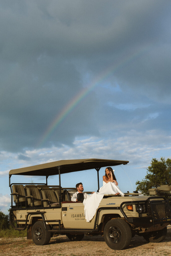 Newlywed couple sitting on a safari vehicle beneath a rainbow after a summer thunderstorm