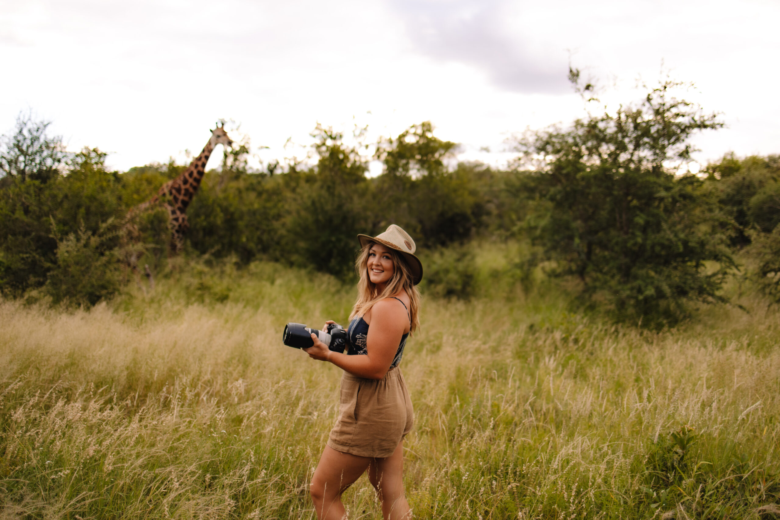 South Africa Safari Elopement photographer Heidi Straus standing in the bushveld holding her camera with a giraffe walking in the background