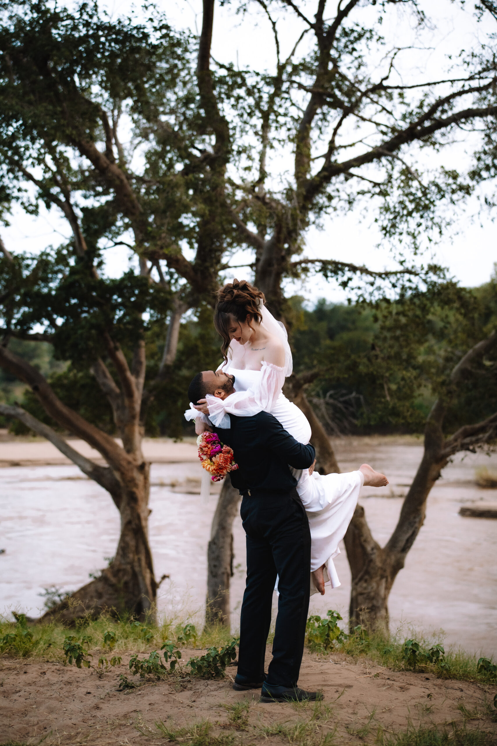 Cape Town elopement groom lifting the bride in his arms beneath trees along a riverbank