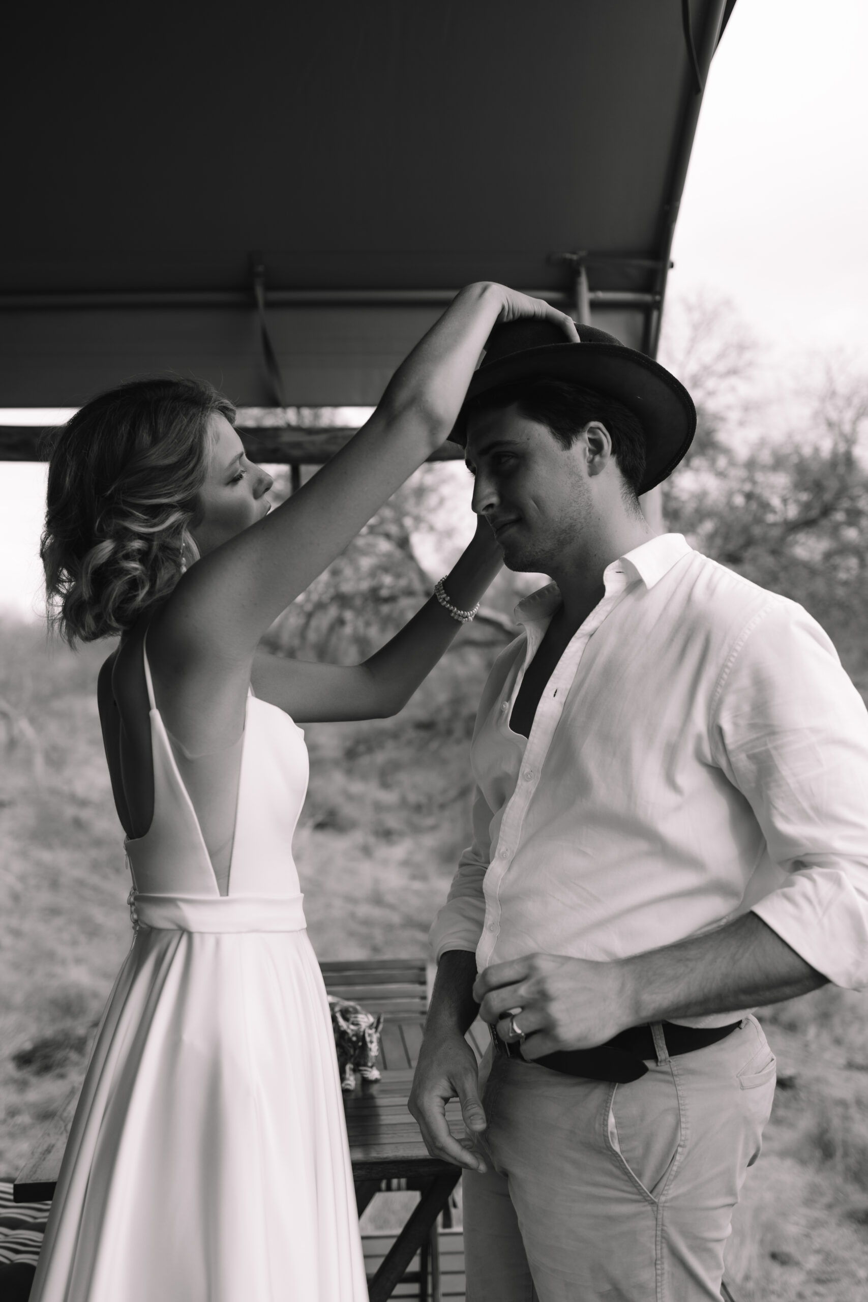 Black and white photo of bride placing a hat on groom during a relaxed safari moment