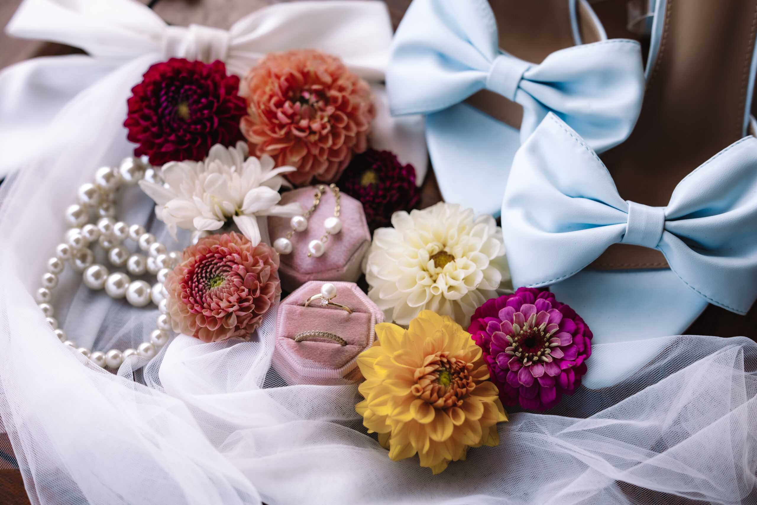 Flat lay of wedding details with colorful flowers, rings, pearls, and blue bow shoes