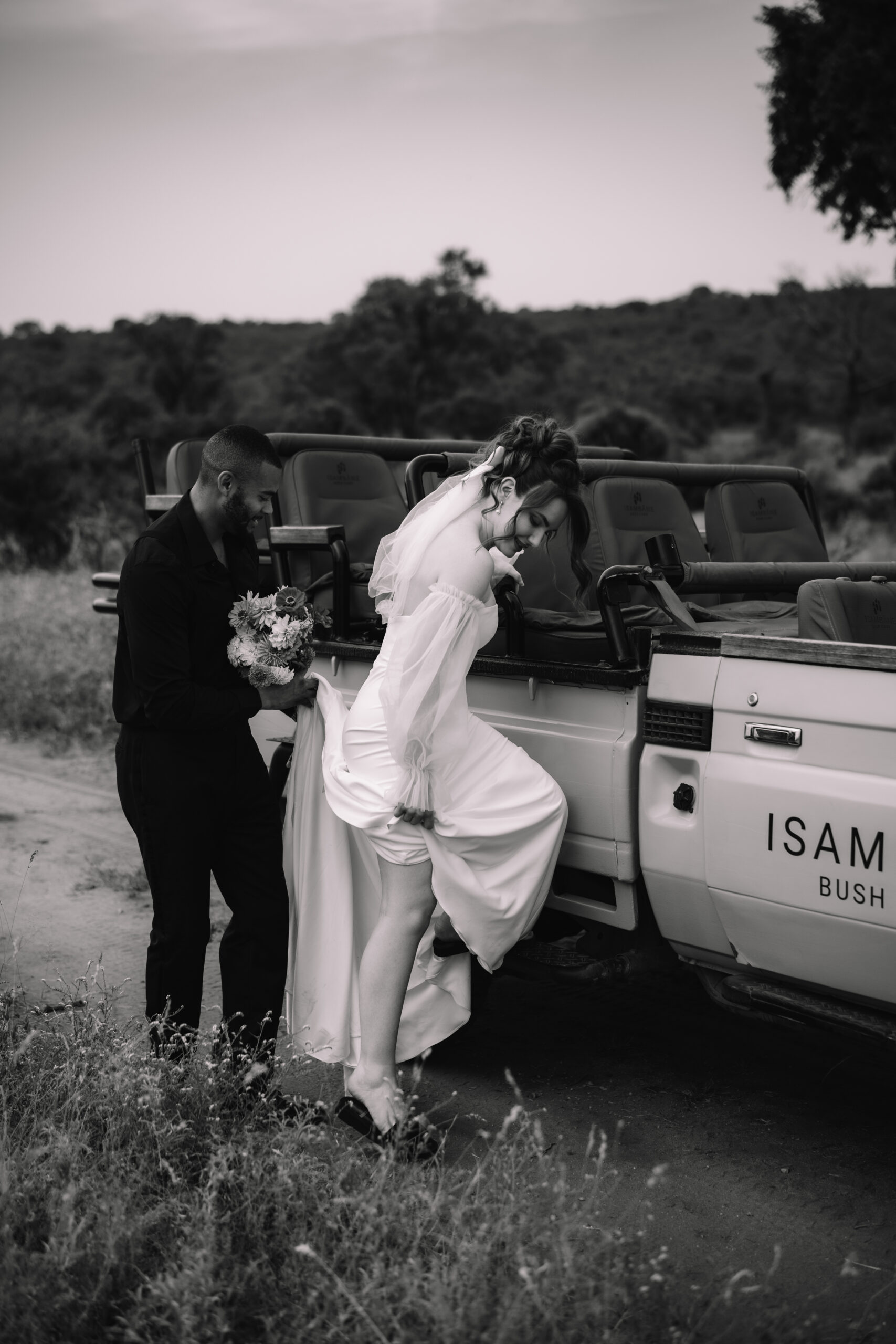 Black and white image of a groom helping his bride climb into a safari vehicle during their wedding day adventure.