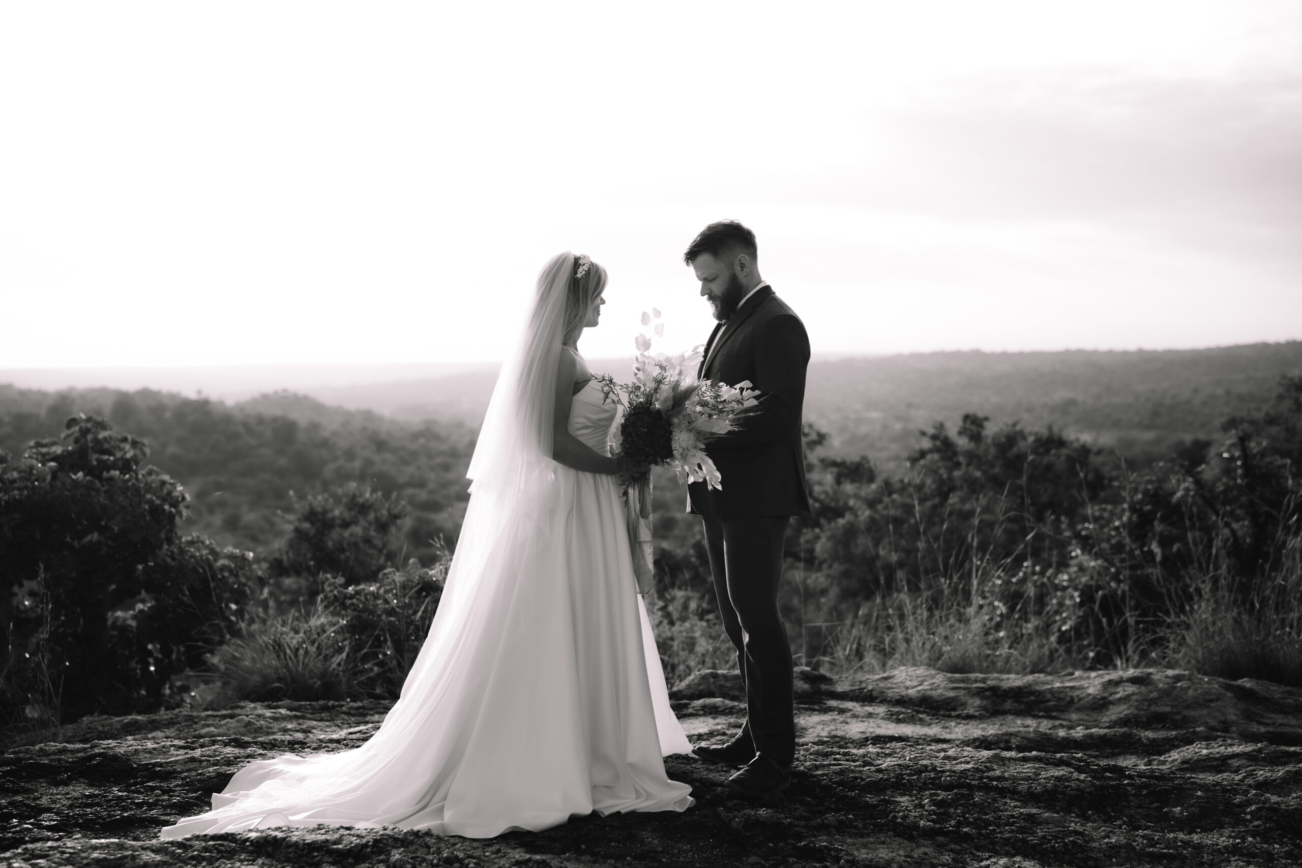Black and white image of a bride and groom facing each other on a rocky overlook with sweeping views of a Private Game Reserve in South Africa.