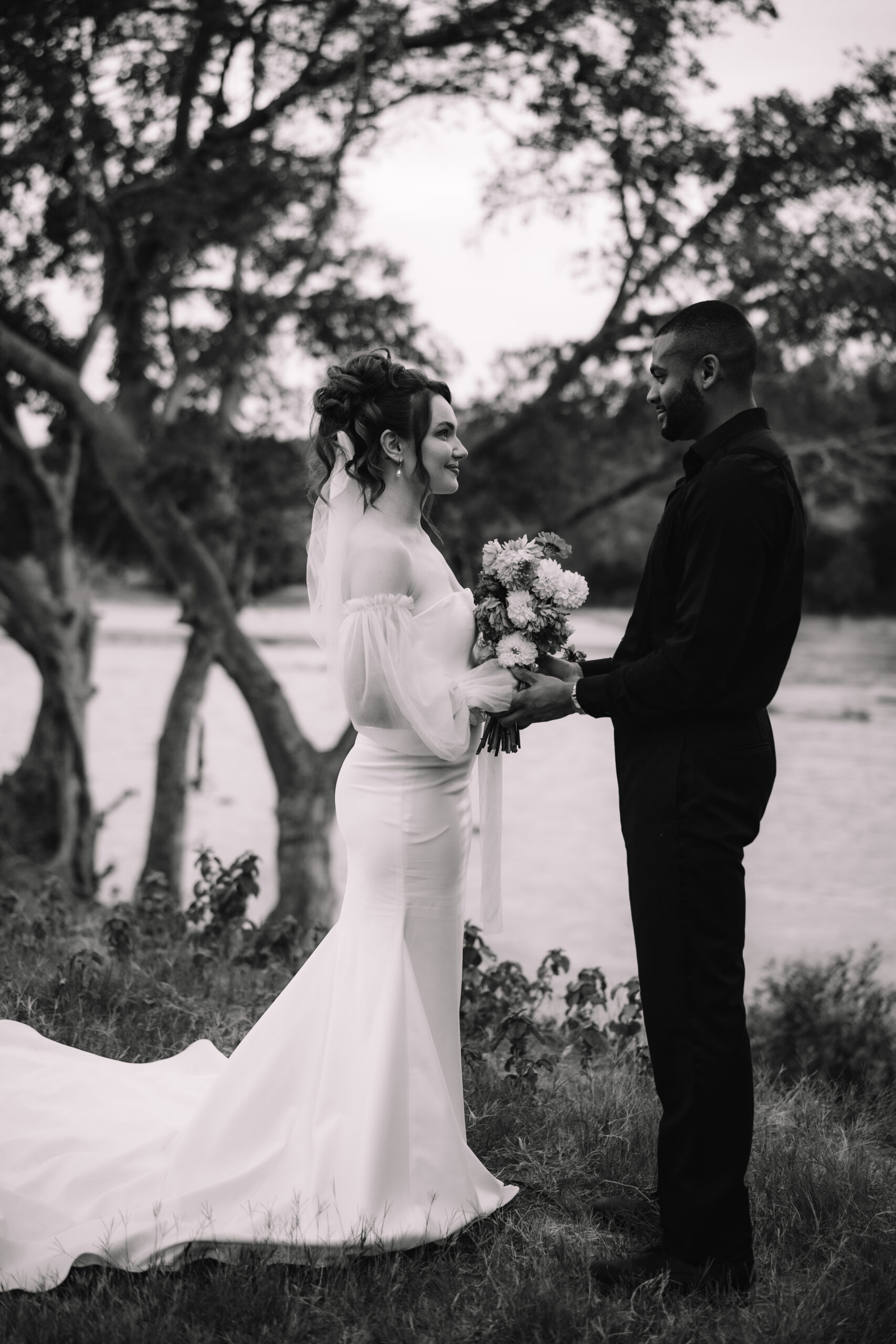Black and white image of a bride and groom exchanging flowers during an intimate riverside wedding ceremony.