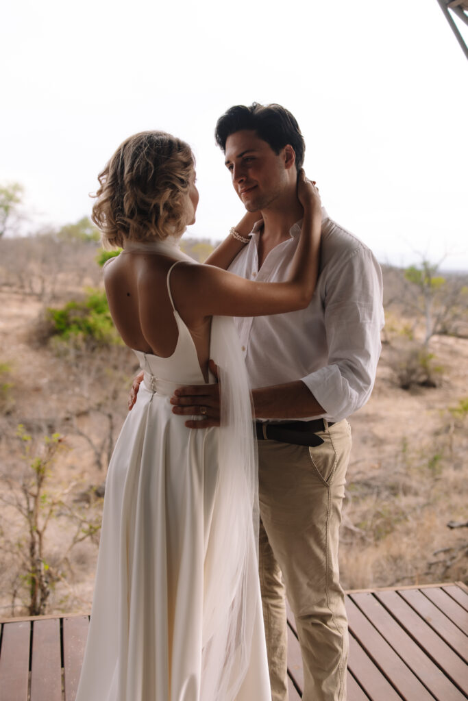 Bride and groom embracing on a wooden deck, surrounded by African bushland during a quiet moment on their wedding day.