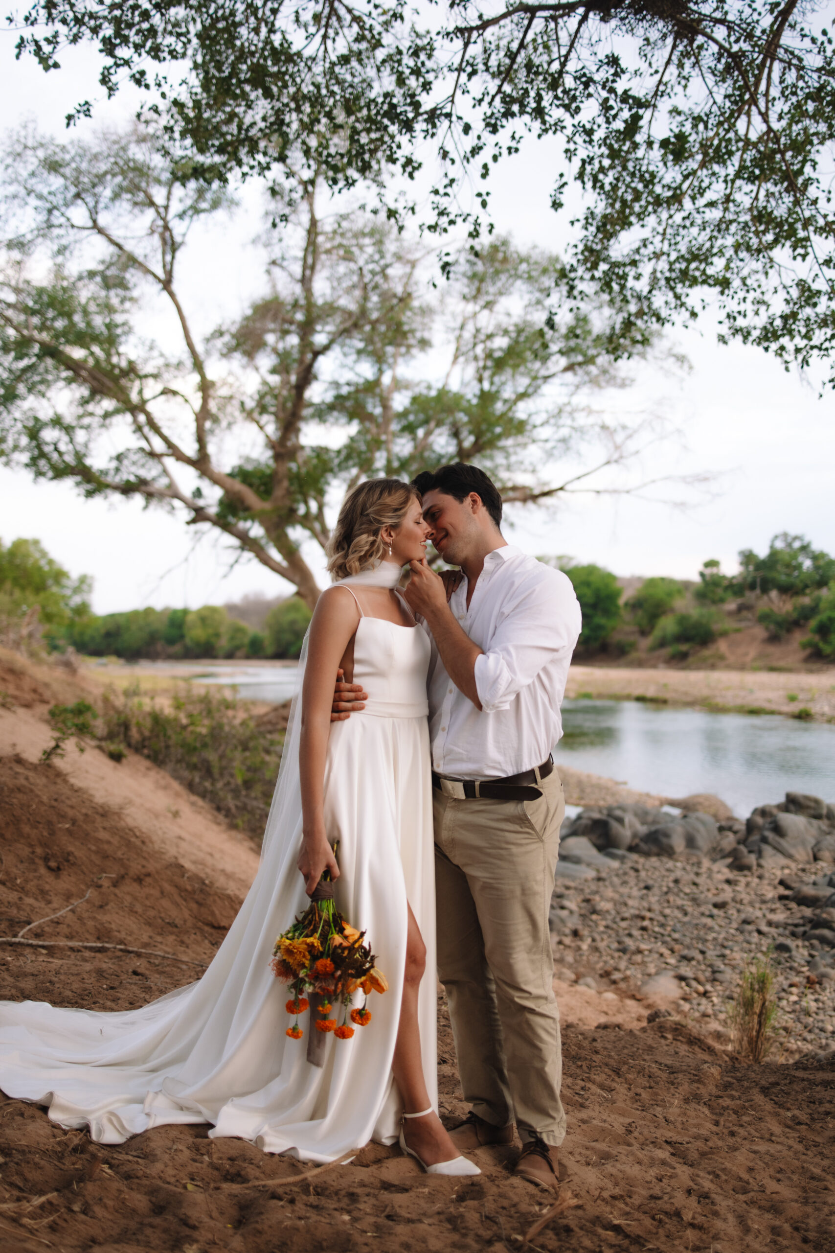 Bride and groom embracing along a riverbank, surrounded by trees and natural landscapes on their wedding day in South Africa.
