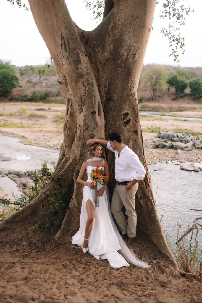 Bride and groom standing beneath a large tree along a riverbank, sharing a romantic moment during their South Africa wedding portraits.
