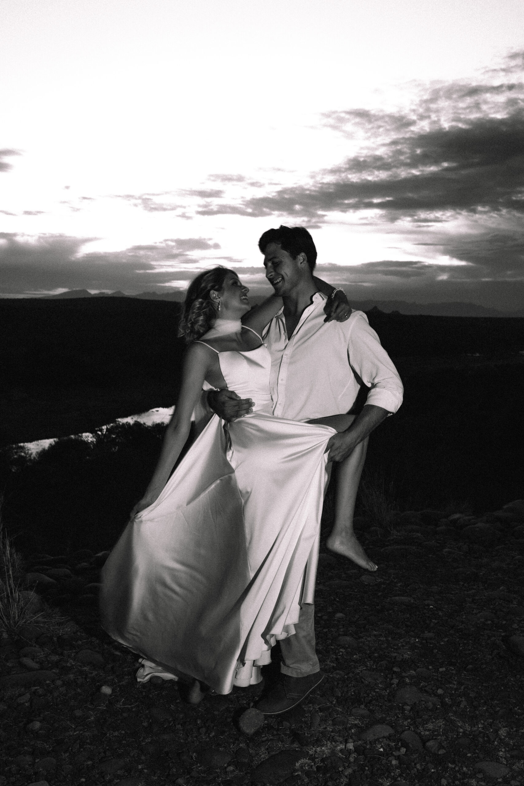 Black and white image of a bride and groom laughing together at sunset on a rocky overlook during their elopement in a Private Game Reserve in South Africa.
