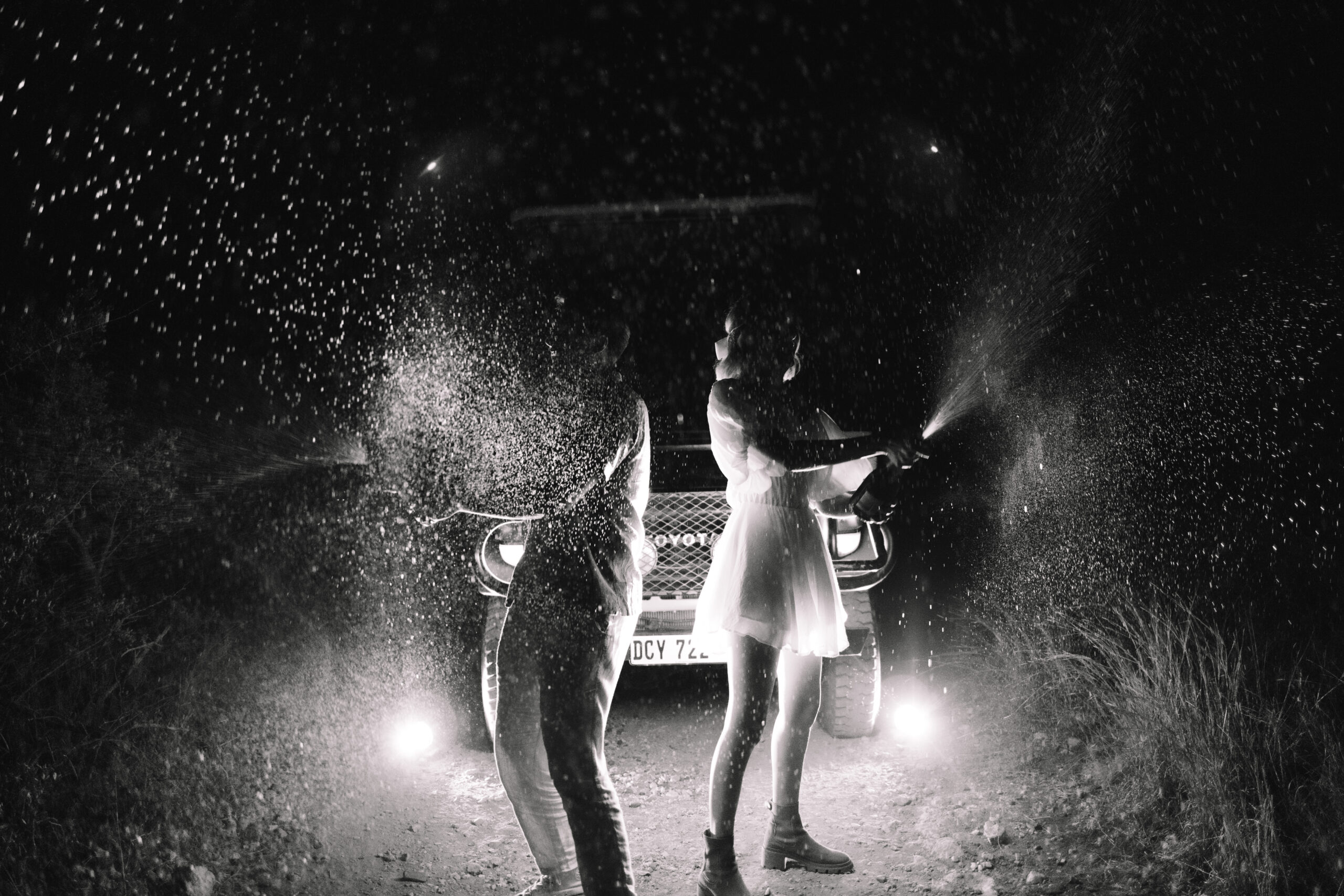 Couple popping champagne at night in front of a safari vehicle, celebrating their elopement under dramatic lighting and flying droplets.