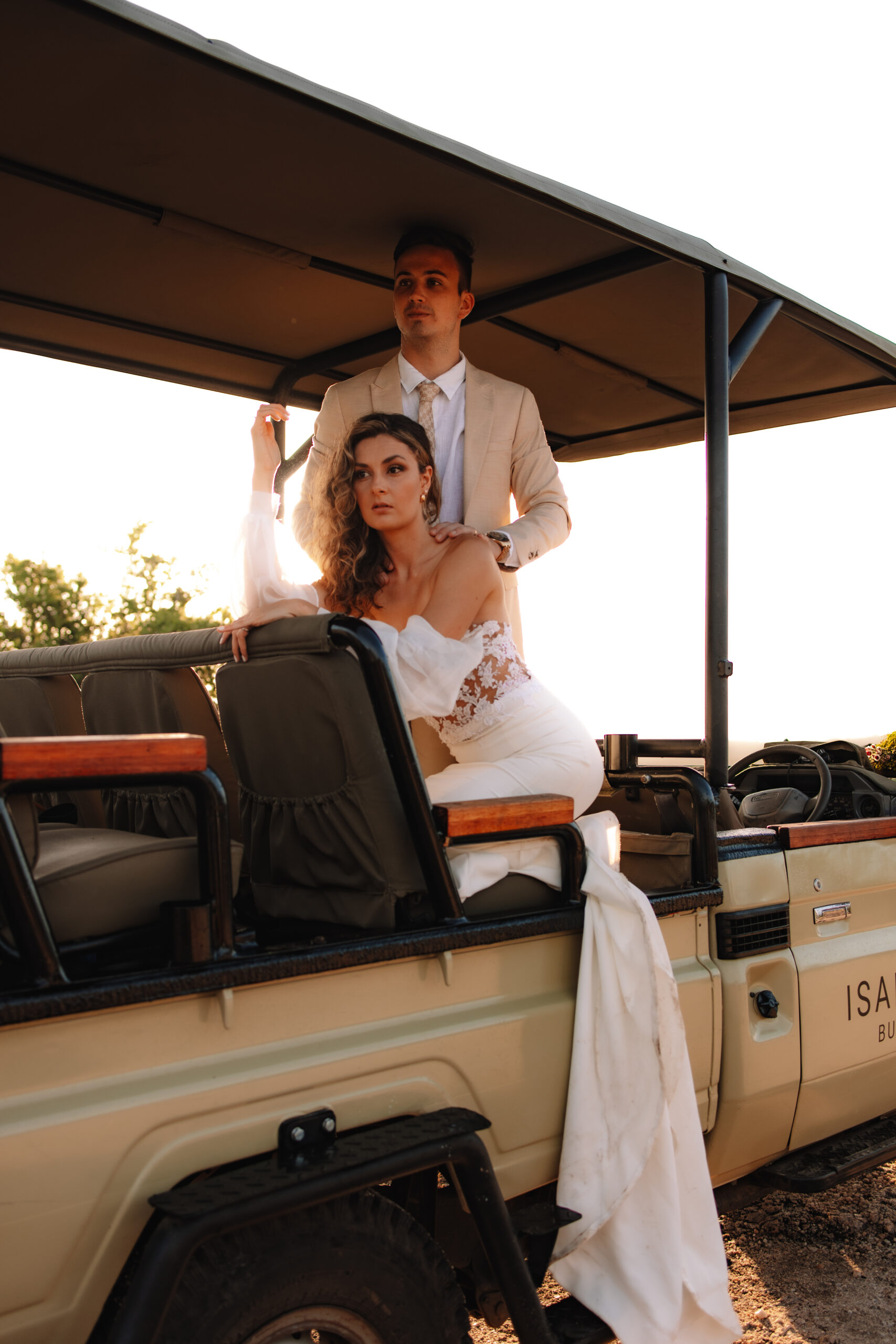 Bride seated on the back of a safari vehicle with her groom standing behind her during sunset wedding portraits.