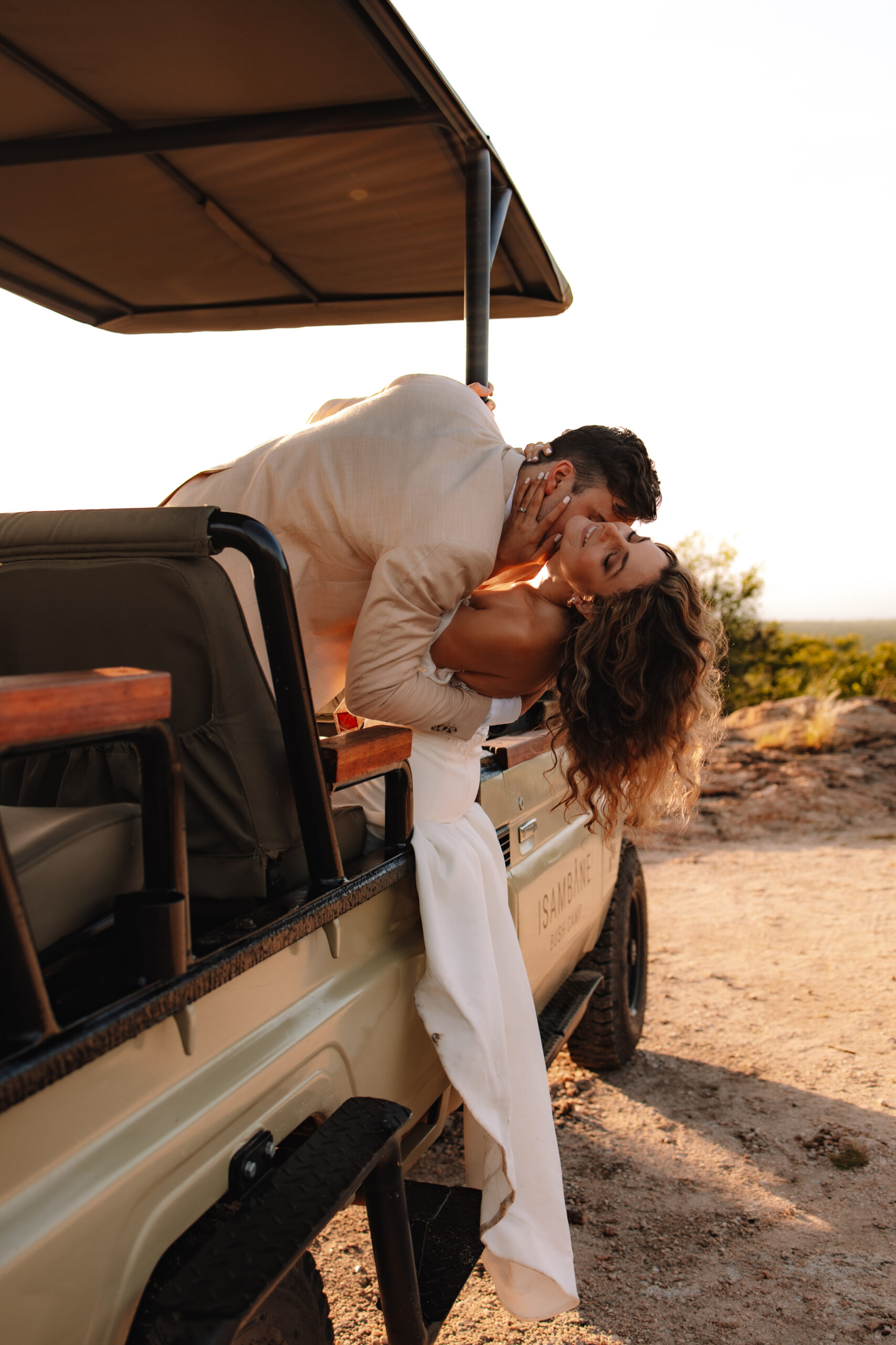 Bride and groom sharing a romantic kiss while leaning out of a safari vehicle during golden hour.