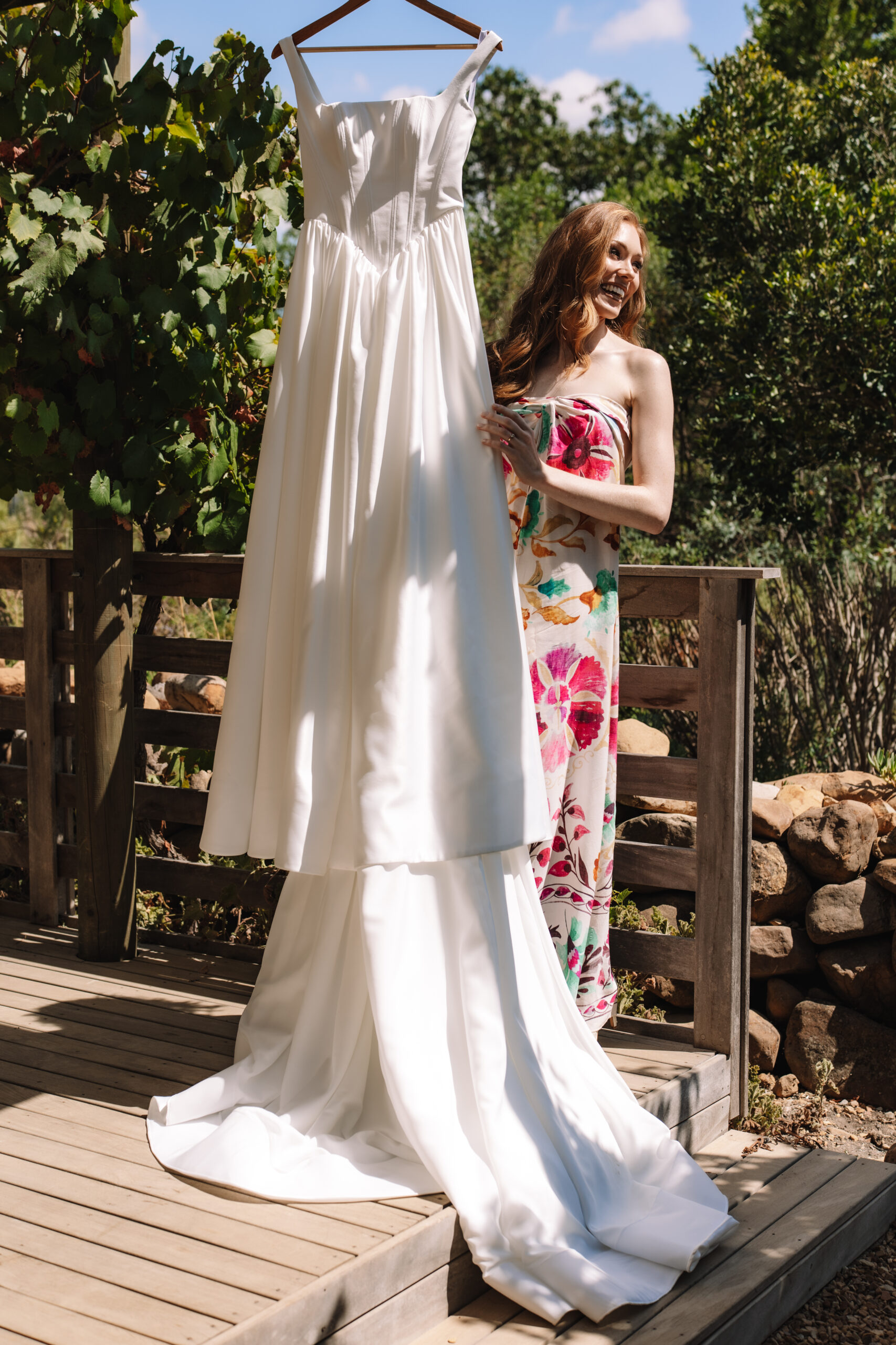 Bride smiling beside her hanging wedding dress on a wooden deck, capturing a quiet getting-ready moment filled with anticipation and light.
