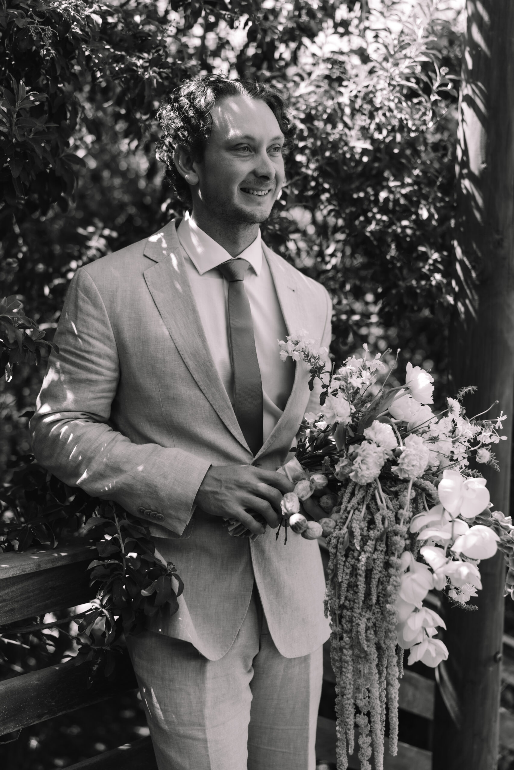 Black and white portrait of a groom holding a cascading bouquet, standing in dappled natural light surrounded by lush foliage.