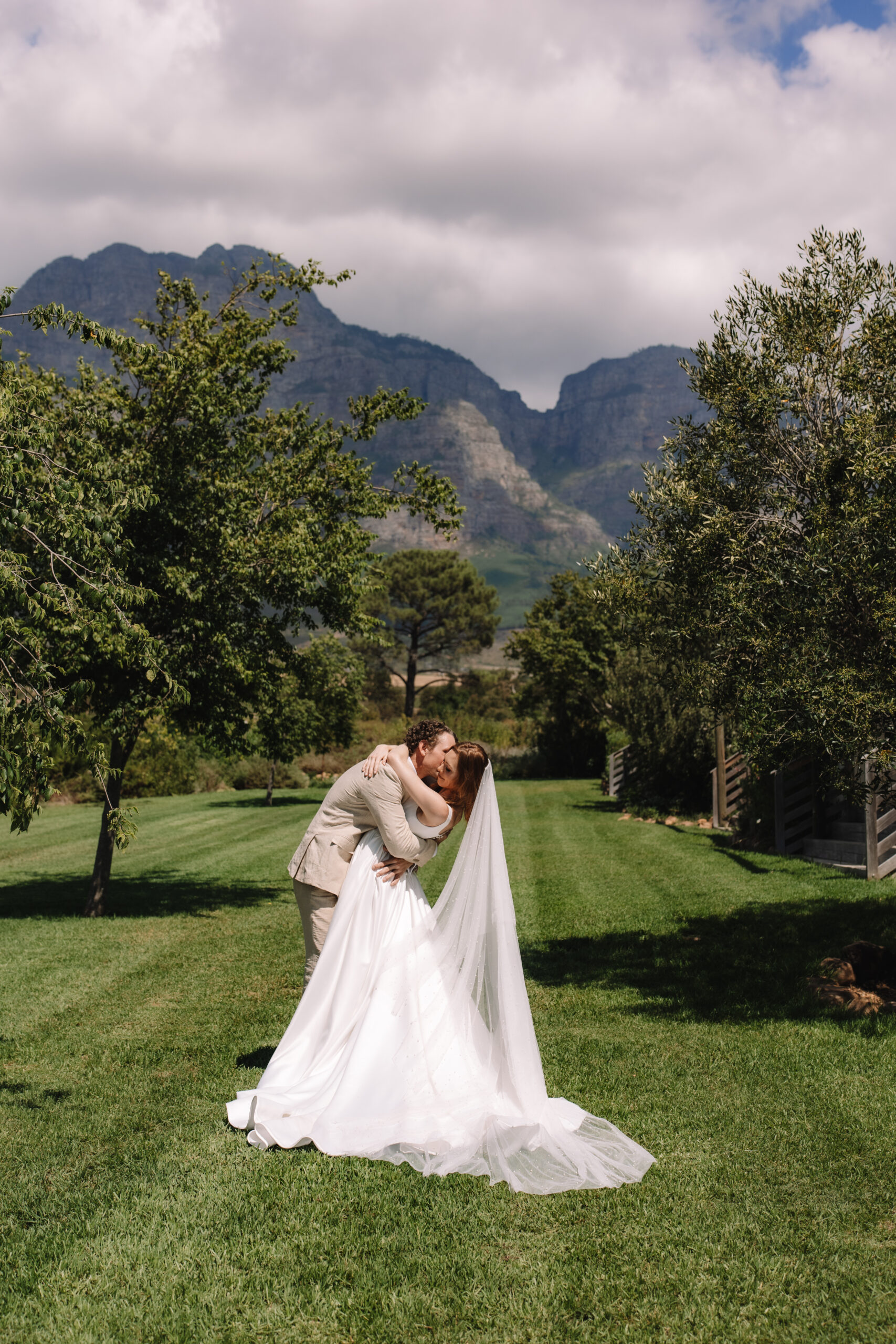 Romantic Cape Town elopement portrait of a bride and groom kissing on a lush green lawn with dramatic mountain views rising in the background.