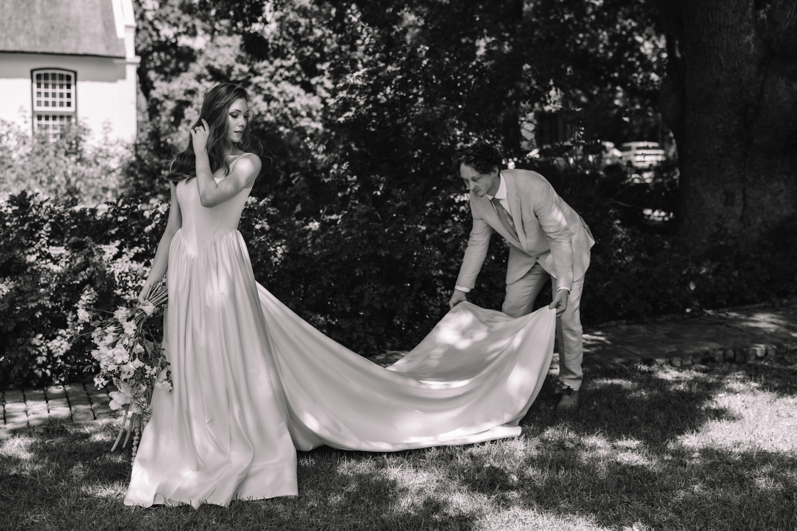 Black and white portrait of a groom gently lifting the train of his bride’s dress during a quiet moment in a garden setting.