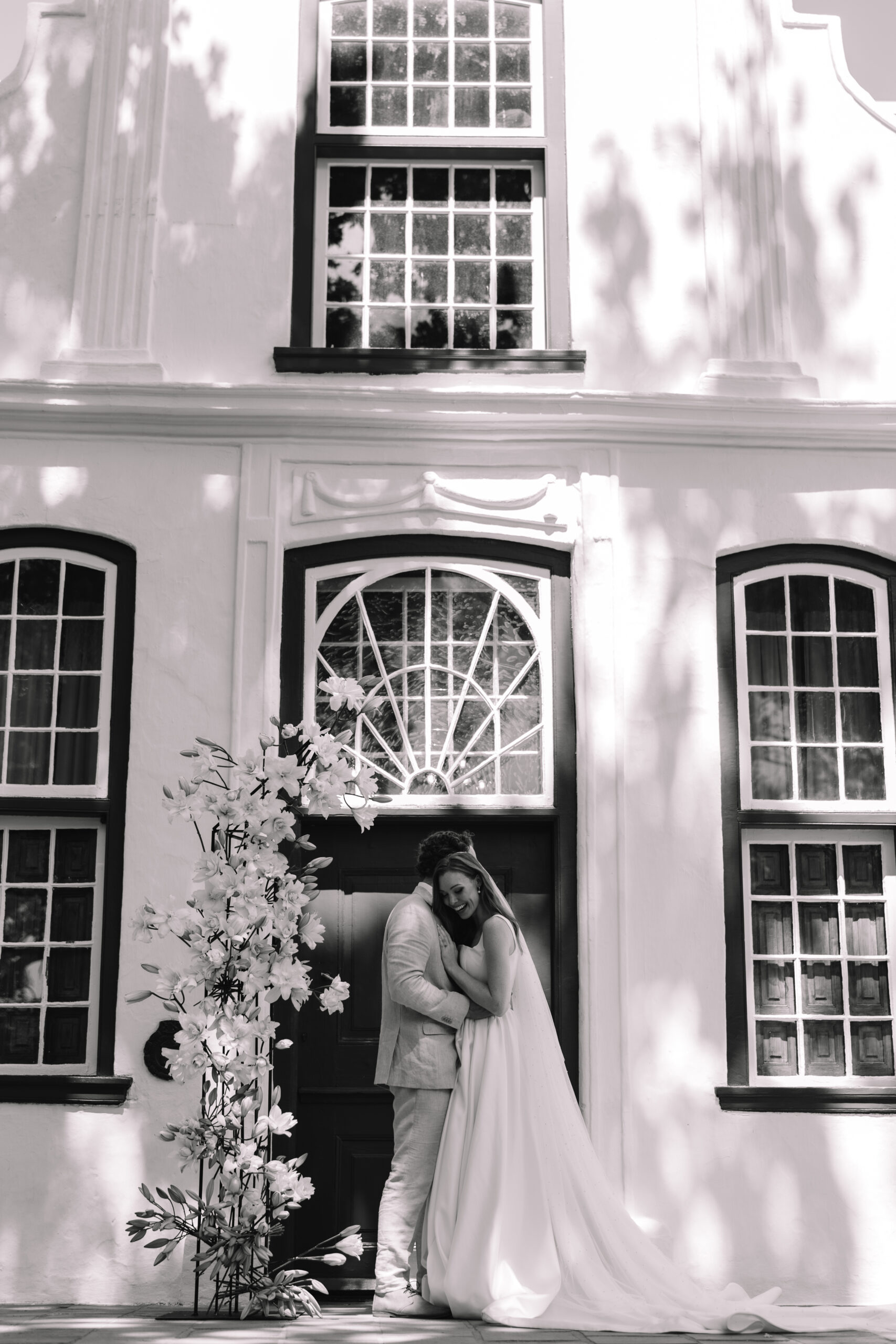 Black and white Cape Town elopement photo of a bride and groom embracing outside a historic white Cape Dutch building, creating a timeless and romantic moment.