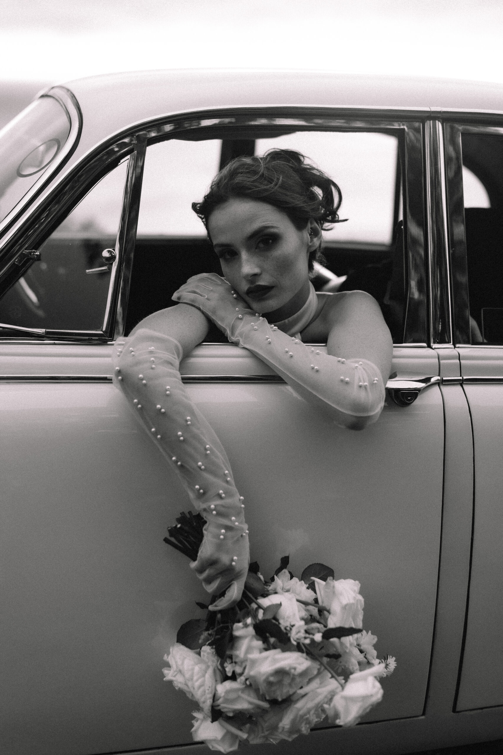 Editorial black and white Cape Town elopement portrait of a bride leaning out of a vintage car window, holding a bouquet with a soft, cinematic expression.