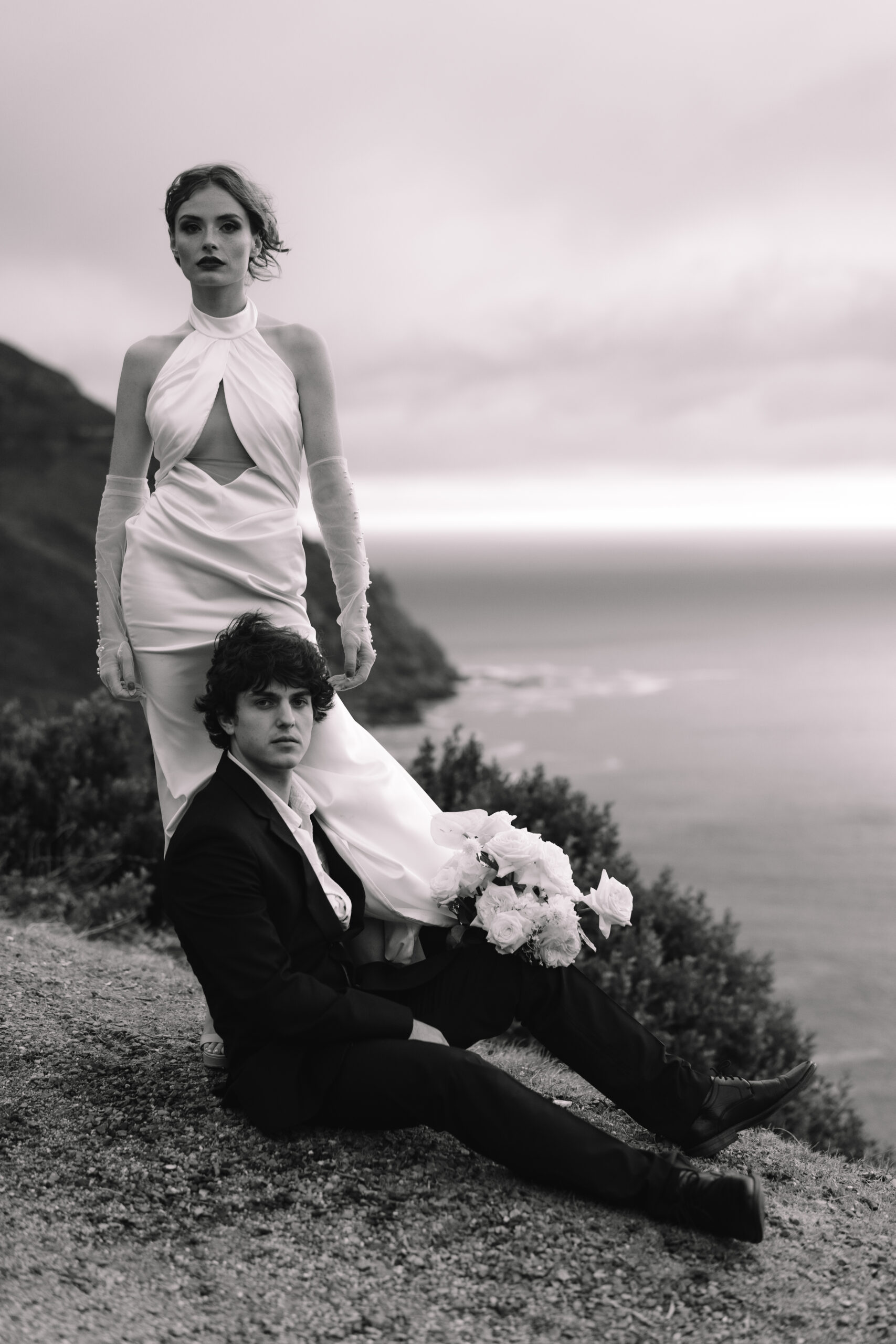 Black and white Cape Town elopement portrait of a bride standing behind her seated groom on a coastal cliff, with dramatic ocean views and wind-swept styling.