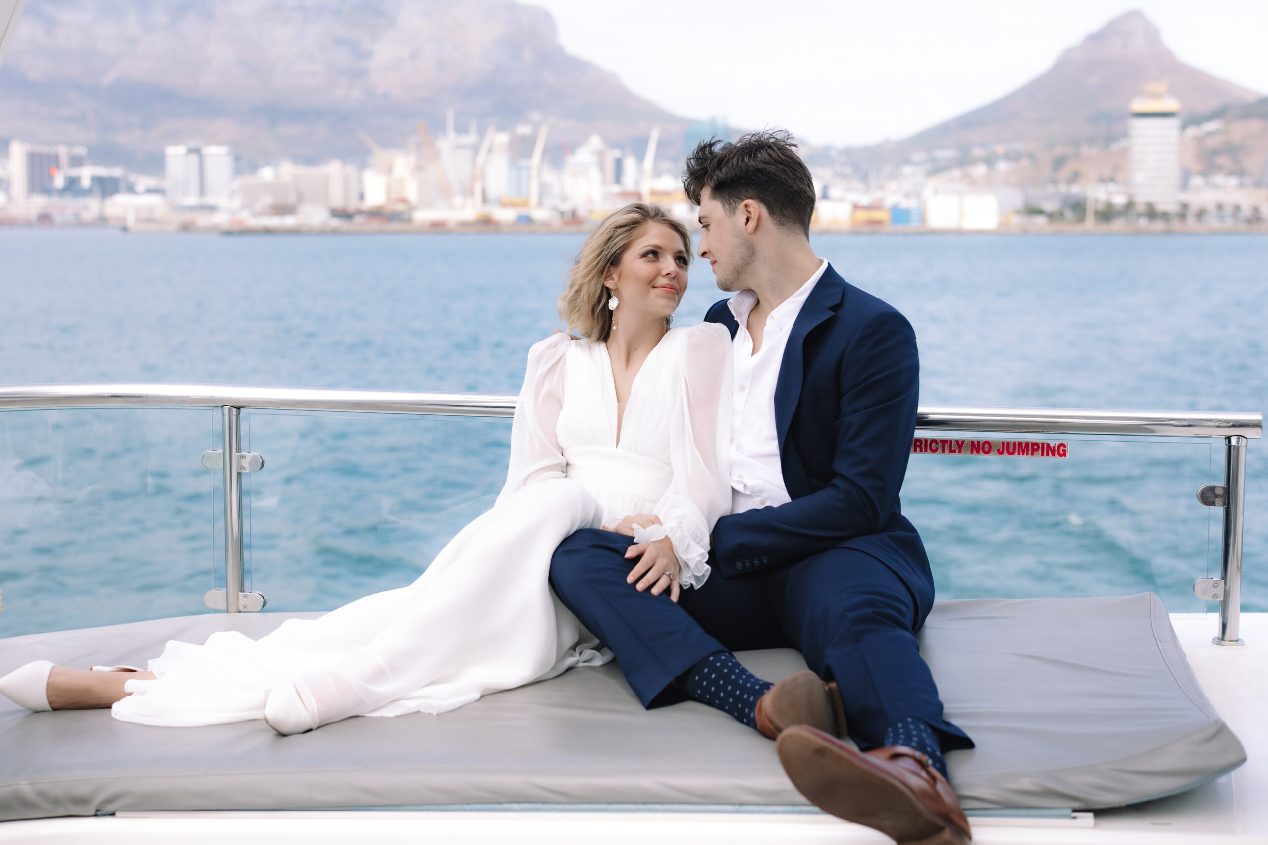 portrait of a newly married couple seated together on a yacht, with harbor views and iconic mountains behind them.