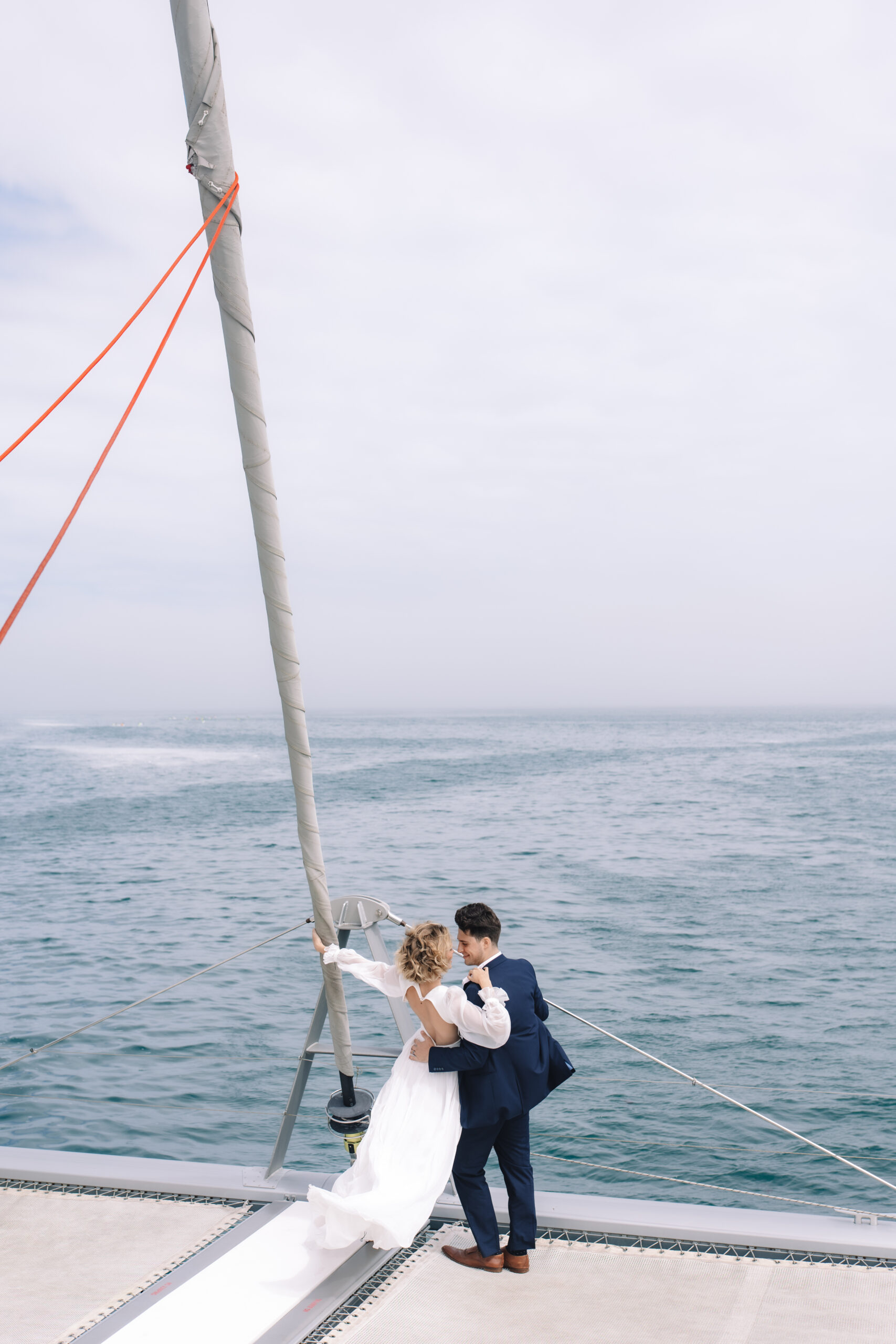 Wide-angle photo of a couple sharing a quiet embrace on a catamaran at sea, surrounded by open ocean and sky.