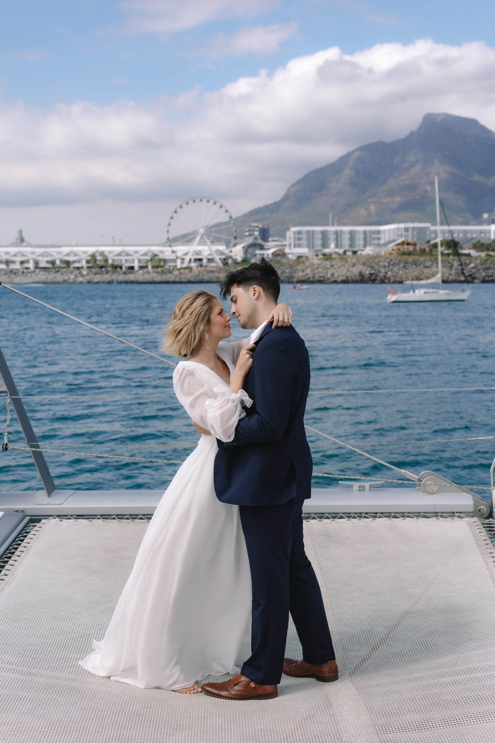 Intimate Cape Town elopement portrait of a bride and groom embracing on a catamaran, with the harbor, ferris wheel, and mountains in the distance.