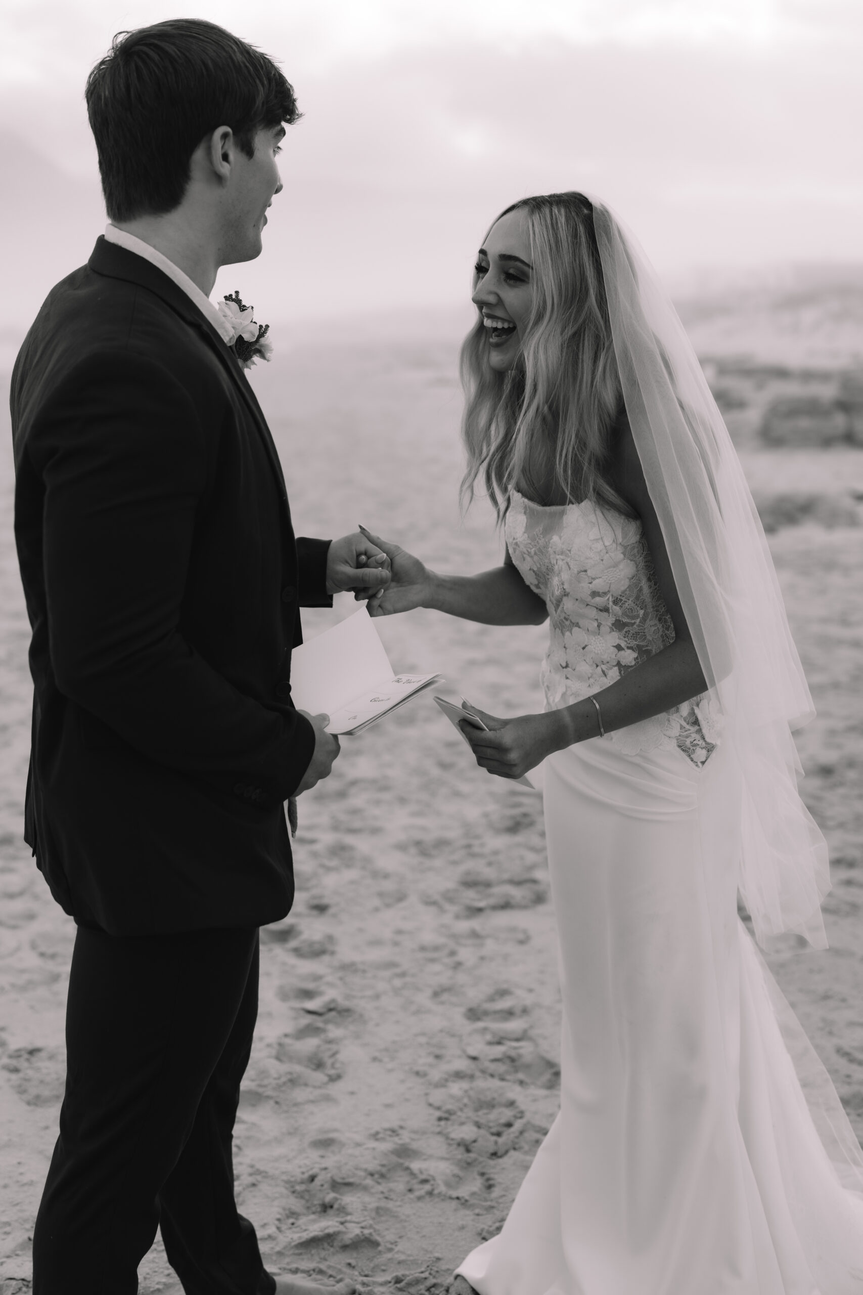 Black and white Cape Town elopement moment of a bride laughing as she reads vows on the beach, holding hands with her groom in an intimate, joy-filled exchange.