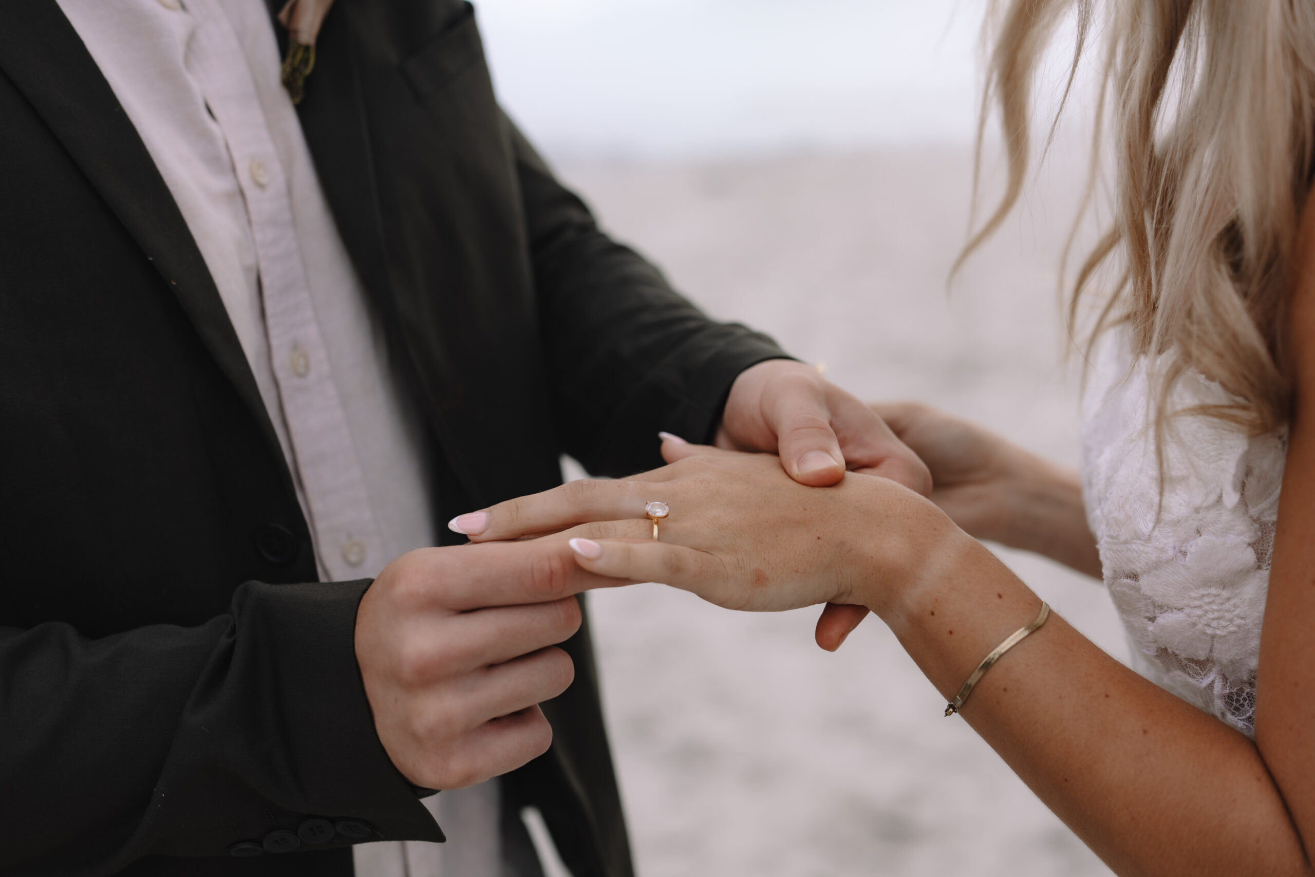Close-up detail of a ring exchange during an intimate beach ceremony, capturing hands, wedding bands, and soft neutral tones.