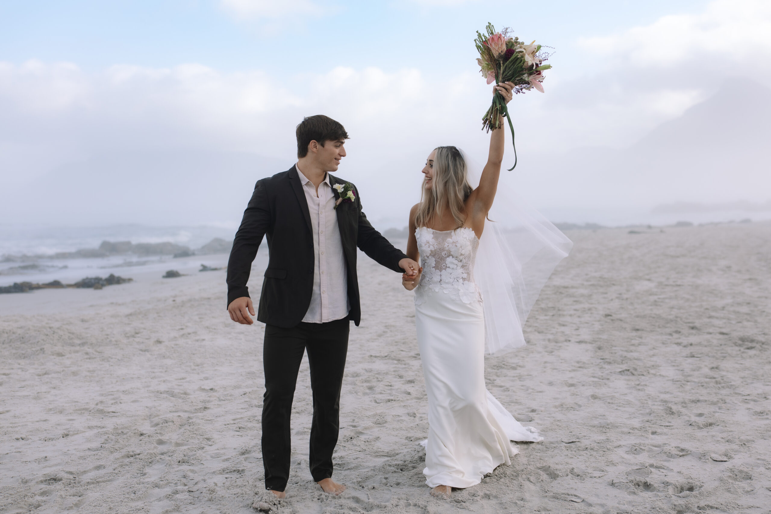 Barefoot couple walking hand in hand on the beach during a Cape Town elopement, bride raising her bouquet in celebration with soft ocean mist and shoreline behind them.
