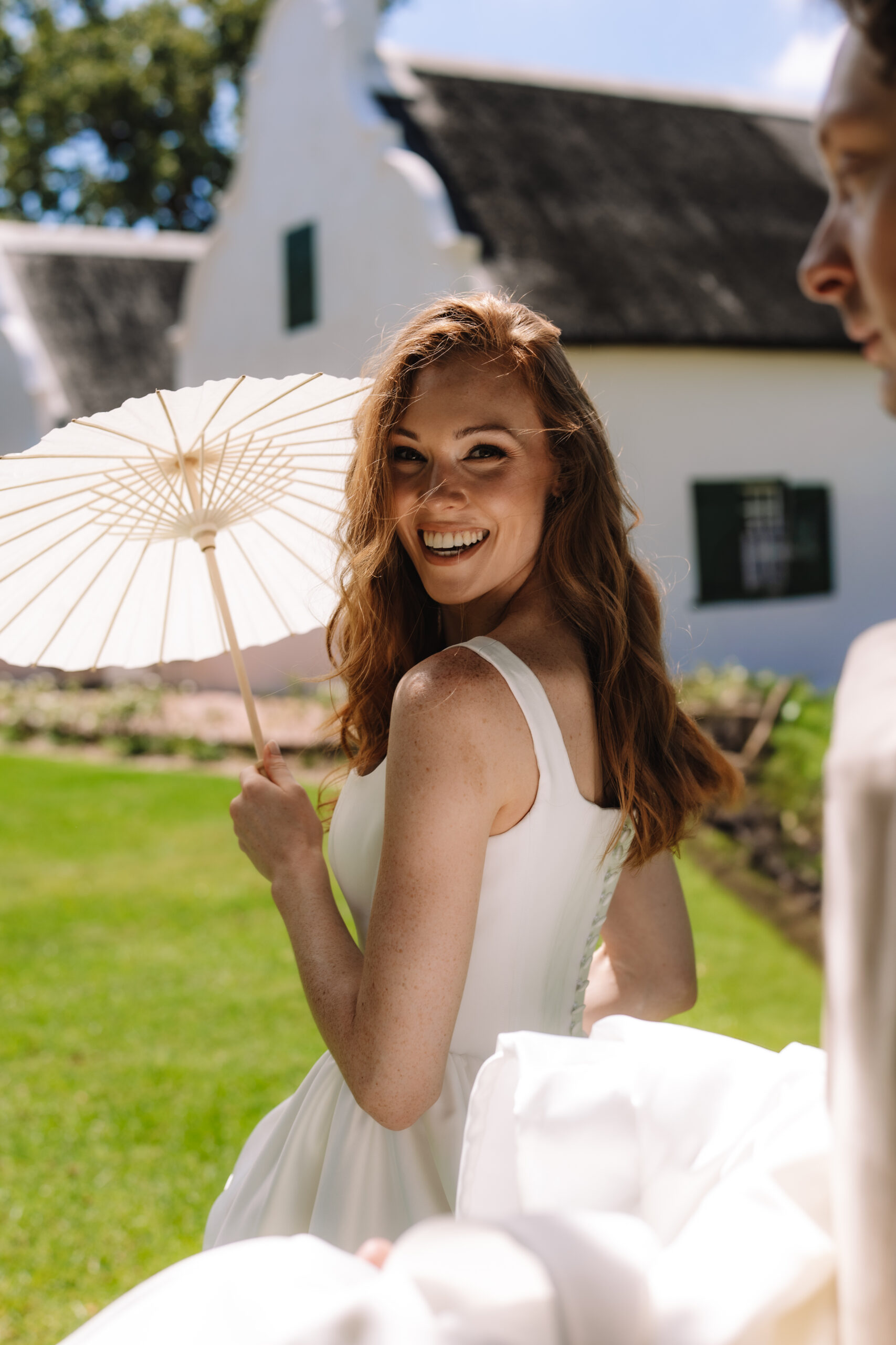 Bride smiling over her shoulder while holding parasol in sunlit garden setting.