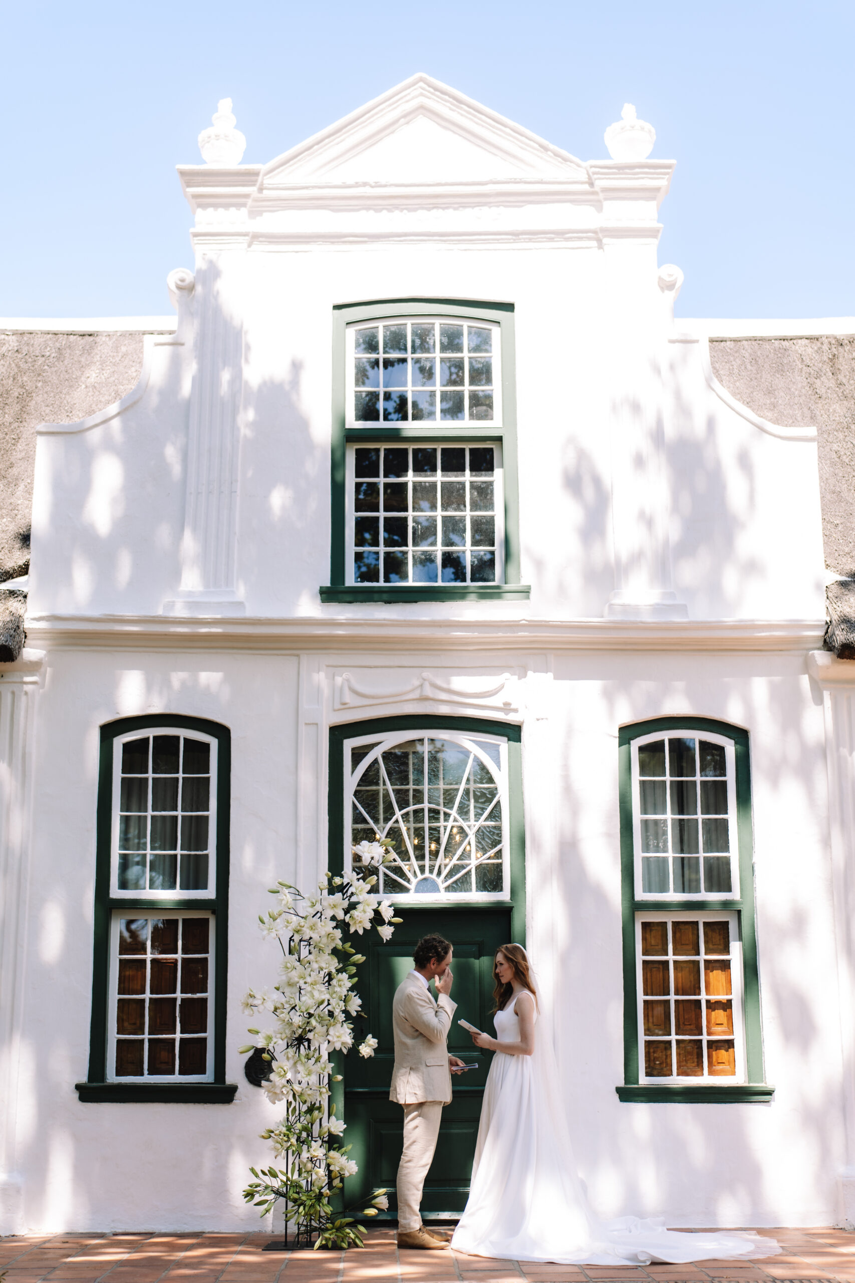 Couple standing beneath white Cape Dutch facade during vineyard elopement near Cape Town.