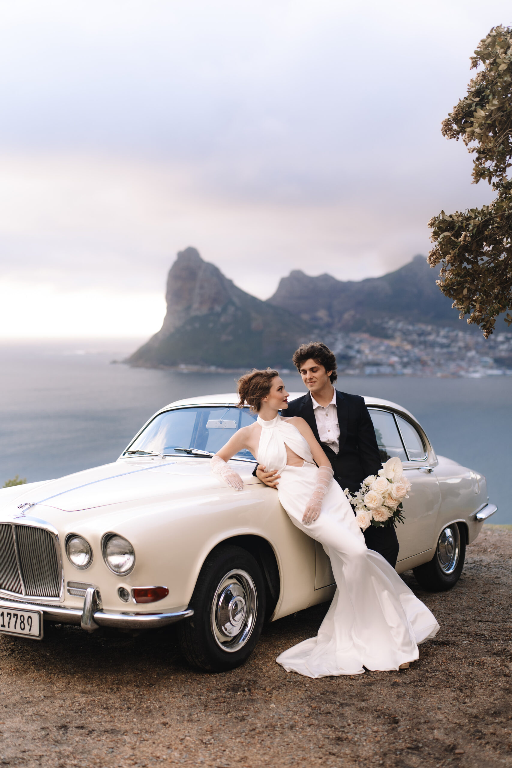 Elegant couple leaning on classic car overlooking coastal cliffs near Cape Town.