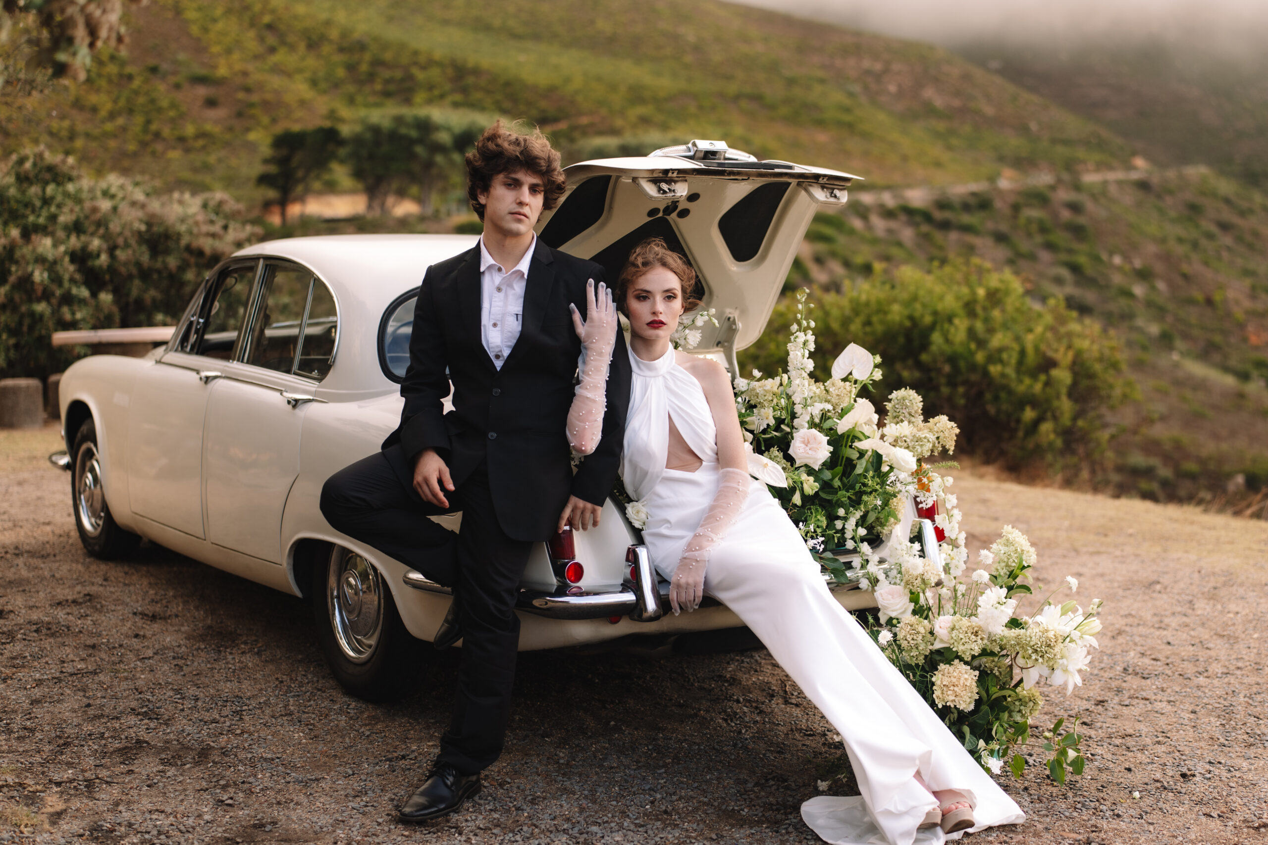 Newlyweds posed with vintage car and floral installation overlooking Western Cape mountains.