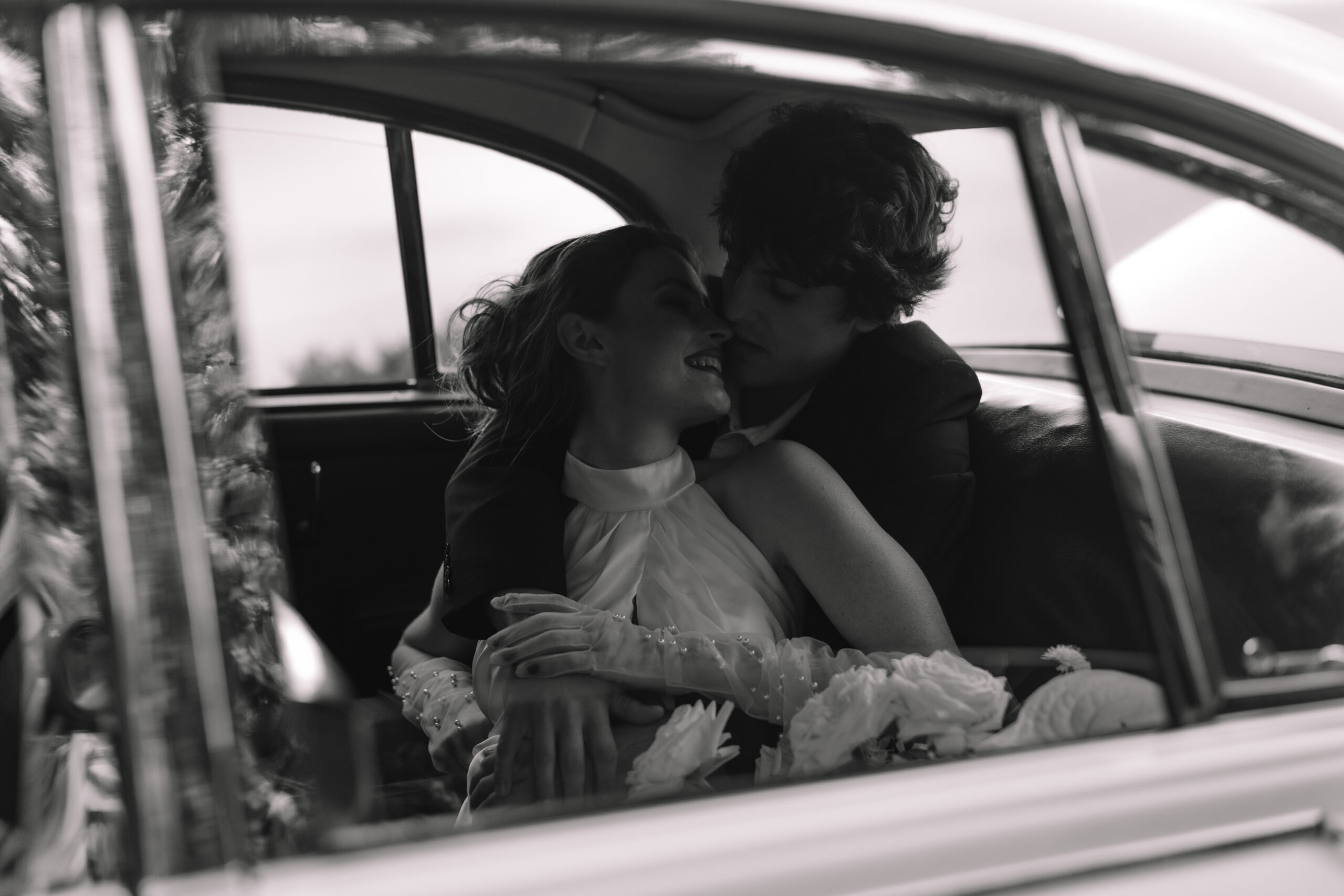 Black and white candid of newlyweds embracing inside vintage car after ceremony.