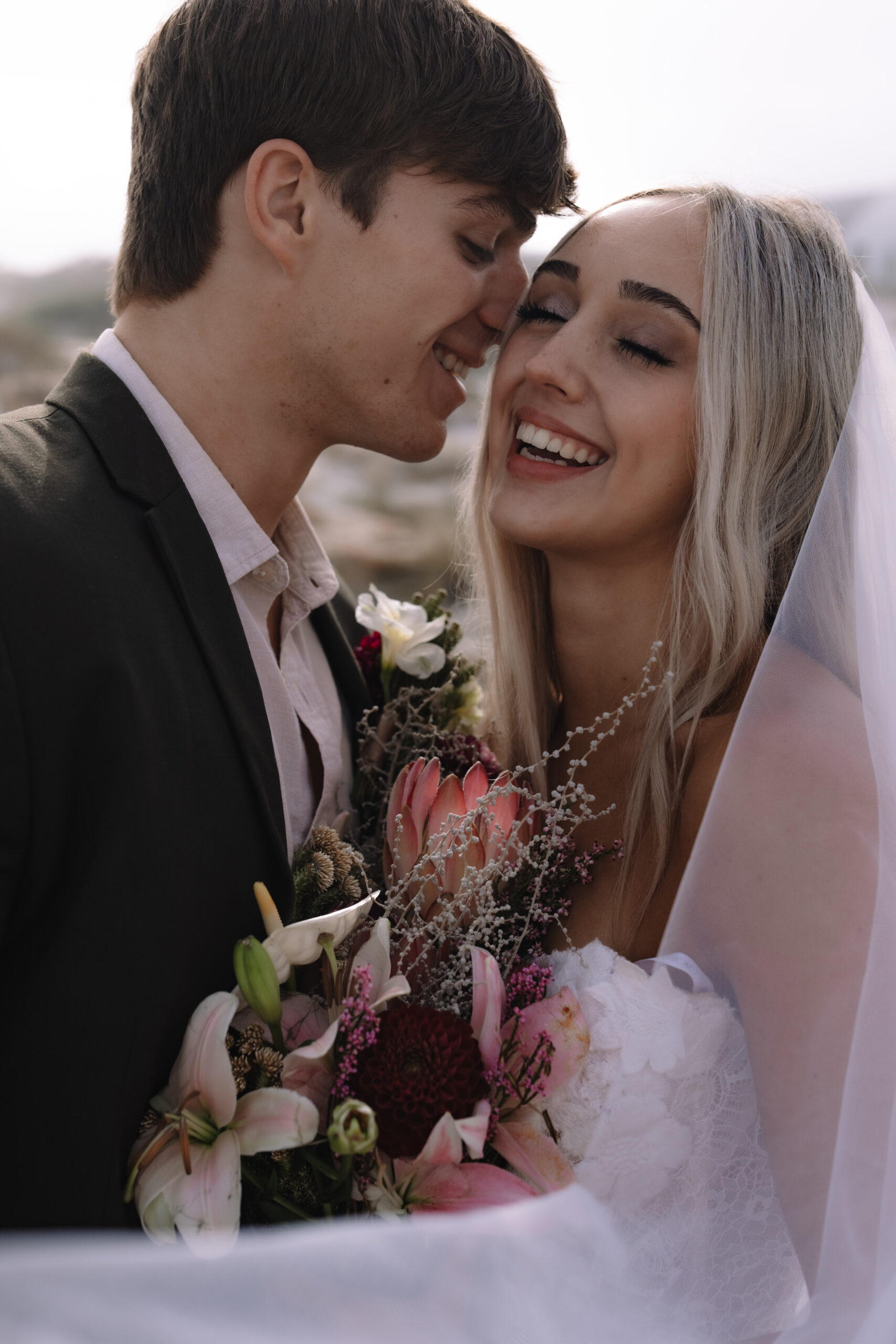 Joyful newlyweds embracing with protea bouquet during seaside wedding celebration.