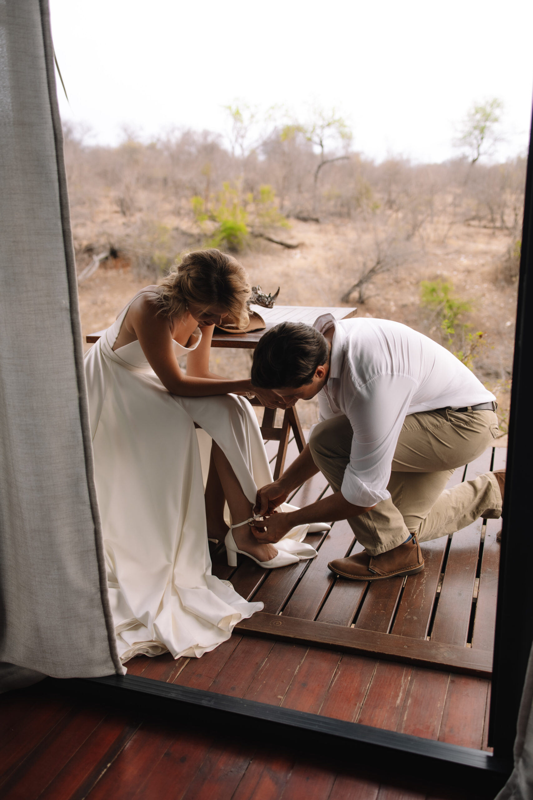 Groom helping bride with her heels on a safari lodge deck during their Cape Town honeymoon.