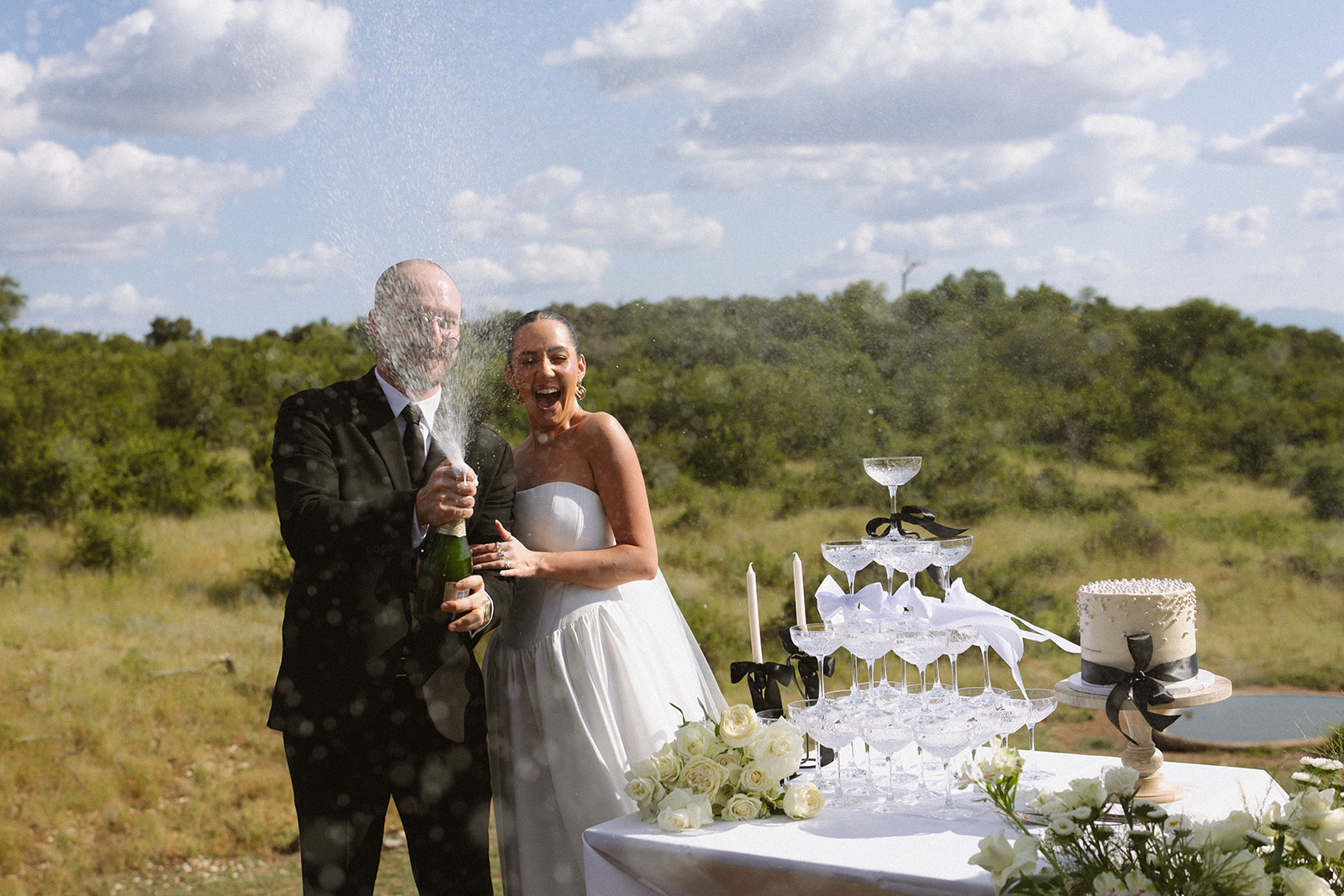 Couple popping champagne beside wedding cake during safari celebration after they elope in South Africa
