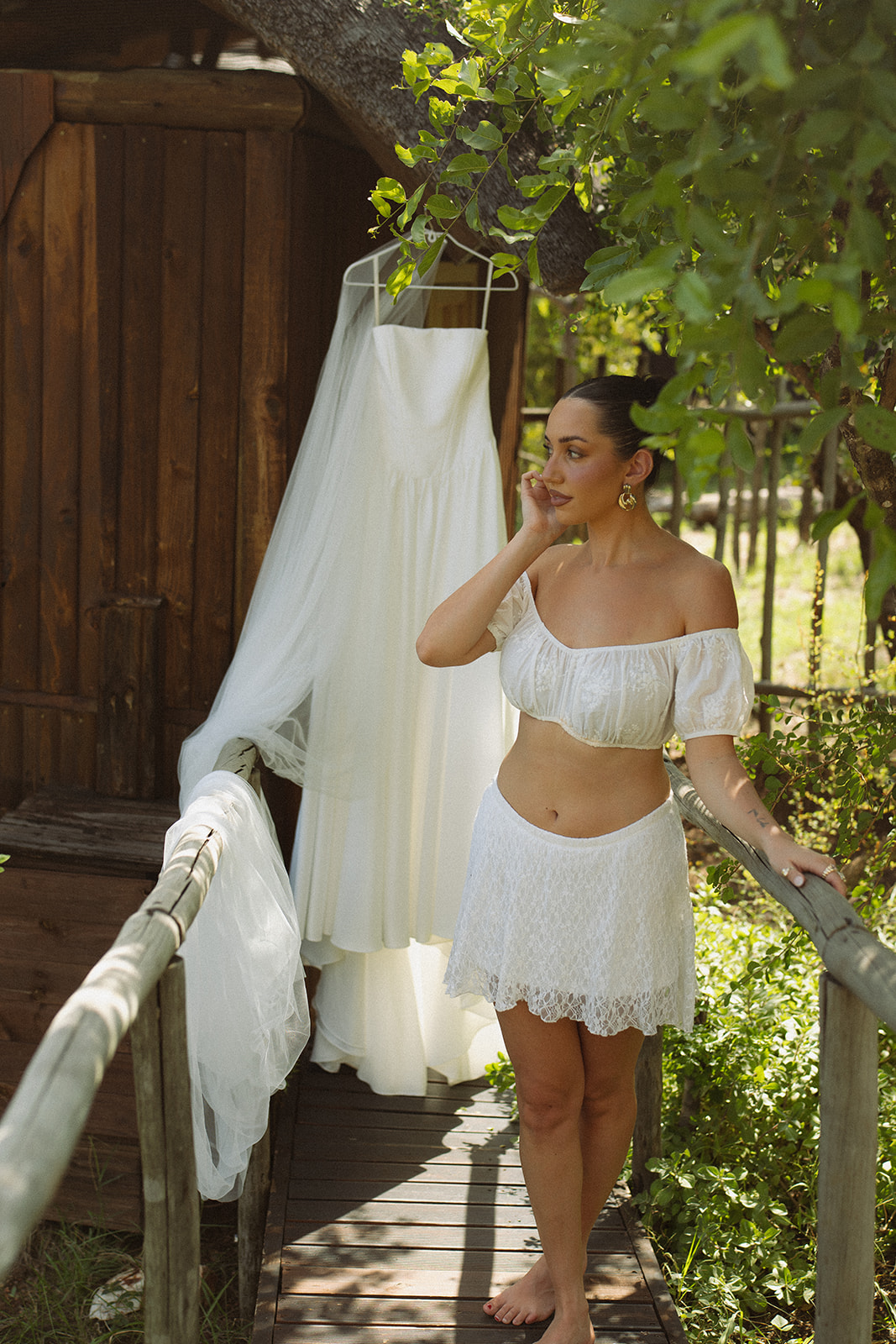 Bride getting ready beside her hanging wedding dress outside a wooden safari lodge surrounded by trees.
