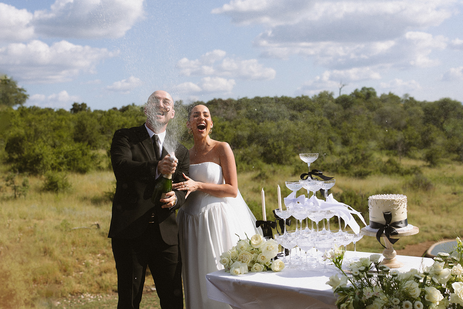 Newlywed couple popping champagne beside a champagne tower and wedding cake during an outdoor safari lodge reception in Africa.
