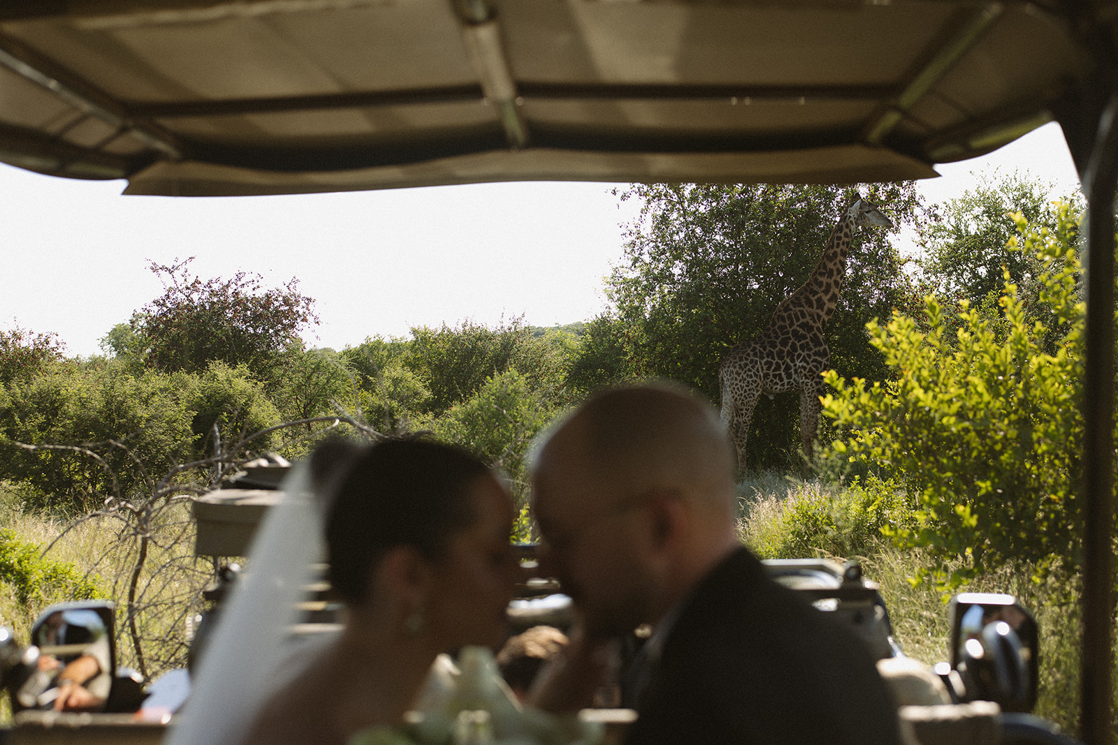 Bride and groom sharing a quiet moment in a safari vehicle while a giraffe stands nearby during their Africa weddings adventure.
