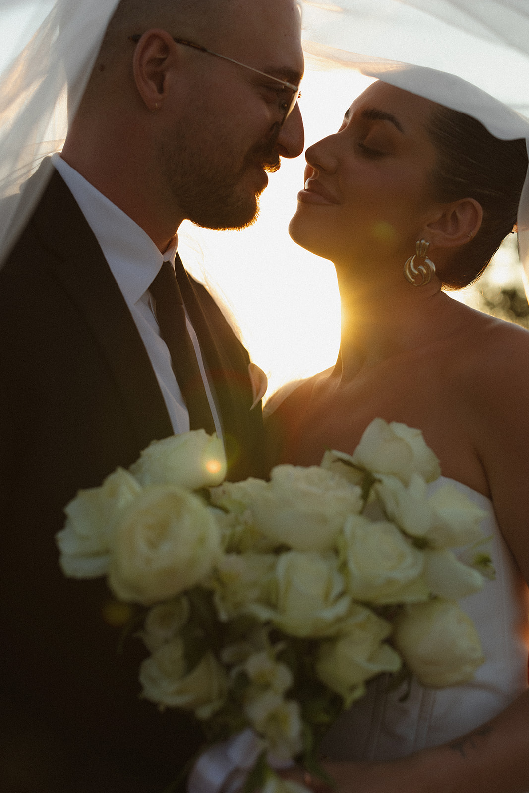 Romantic close-up of bride and groom under a veil at golden hour holding a bouquet of white roses during Africa weddings portraits.
