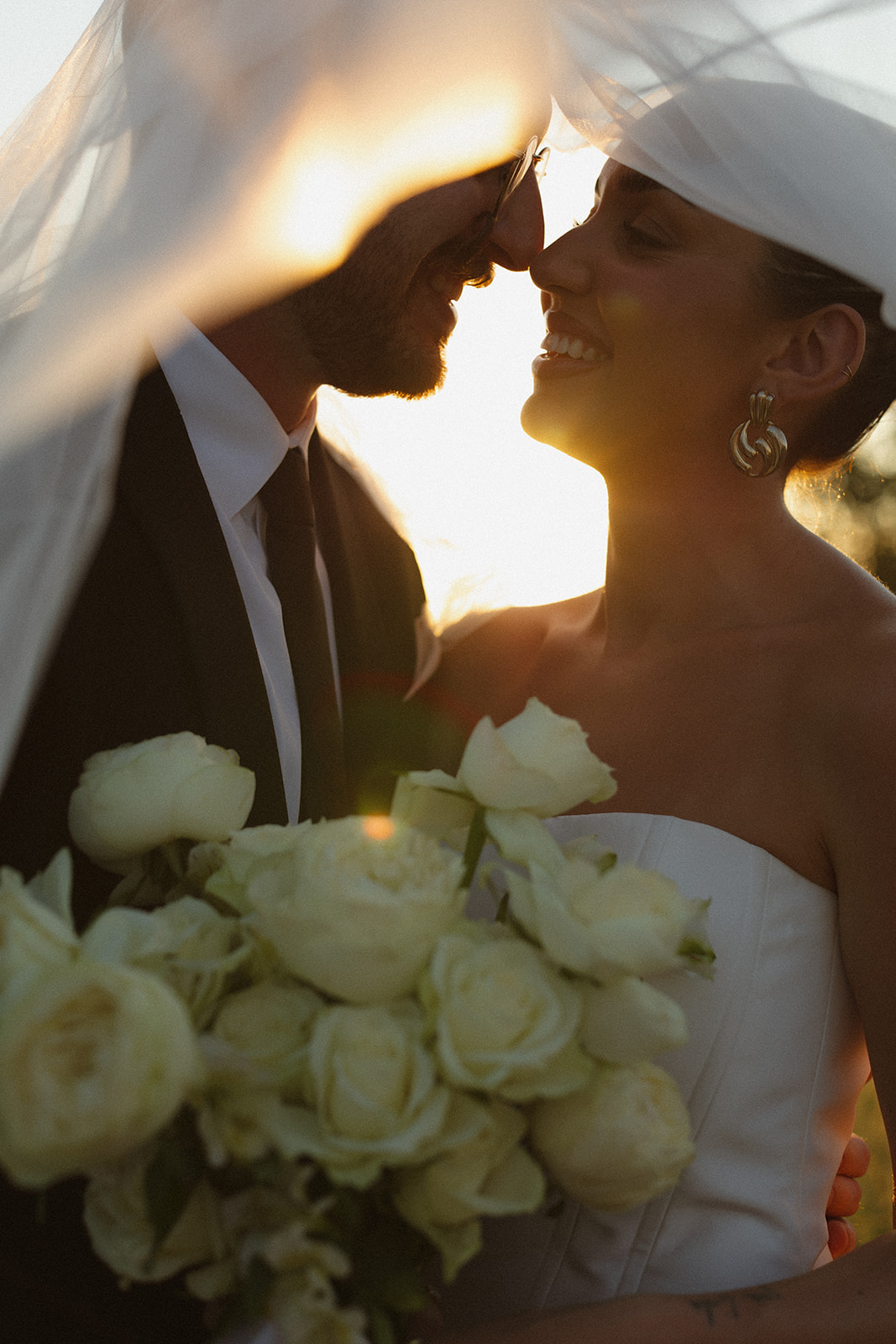 Golden hour portrait of bride and groom smiling under the bride’s veil with a bouquet of white roses during Africa weddings.
