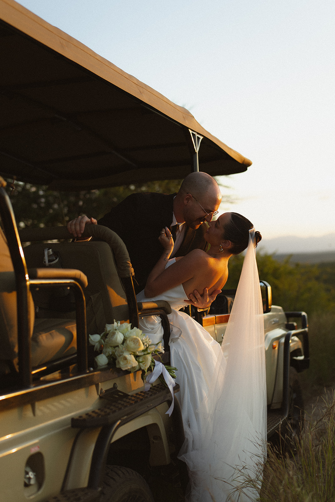 Bride and groom sharing a romantic dip kiss beside a safari vehicle during golden hour portraits for Africa weddings.
