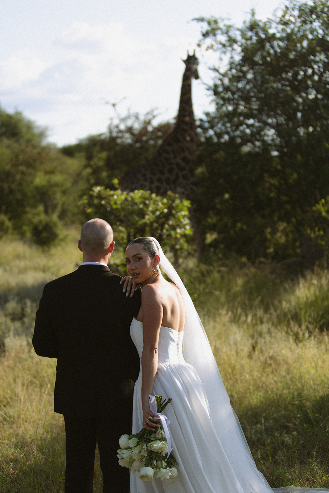 Bride and groom standing in tall grass with a giraffe in the background during a luxury Africa weddings safari experience.
