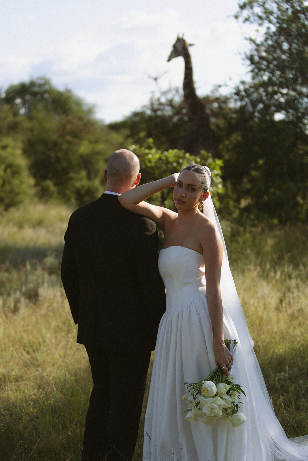 Bride and groom standing in tall grass with a giraffe in the background during romantic portraits for Africa weddings.
