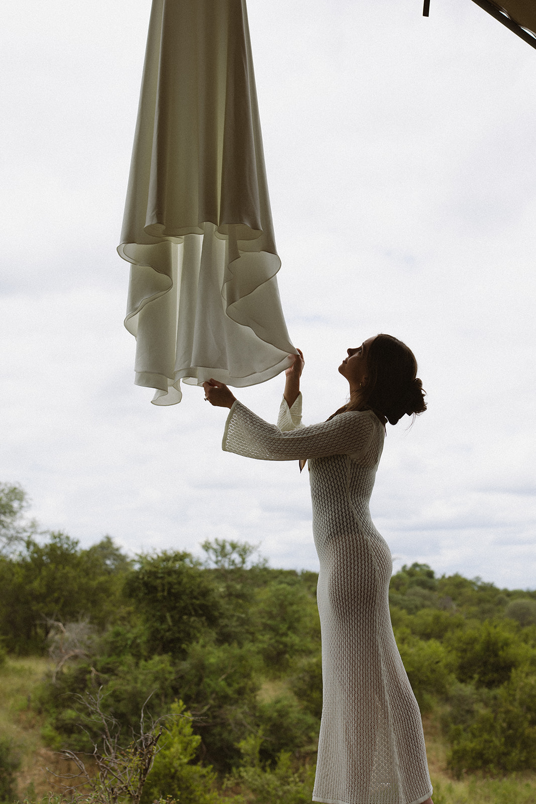 Bride holding her flowing wedding dress outside a safari lodge surrounded by lush greenery
