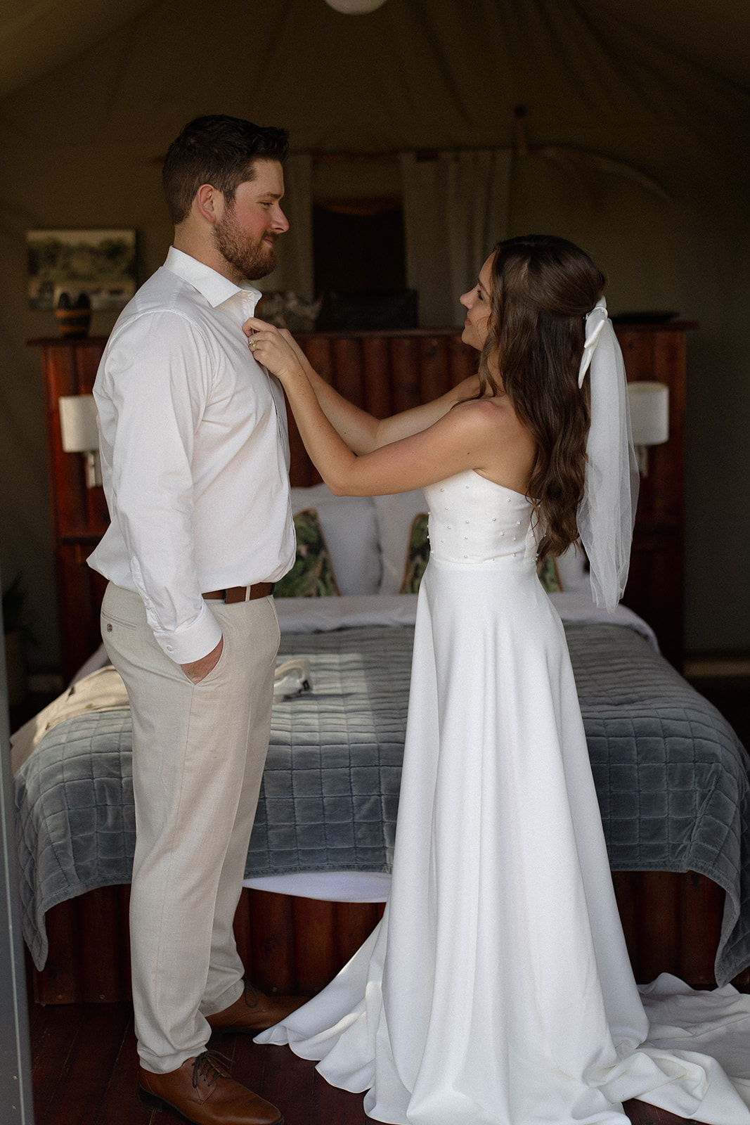 Bride adjusting groom’s shirt inside a safari lodge during a quiet wedding morning moment
