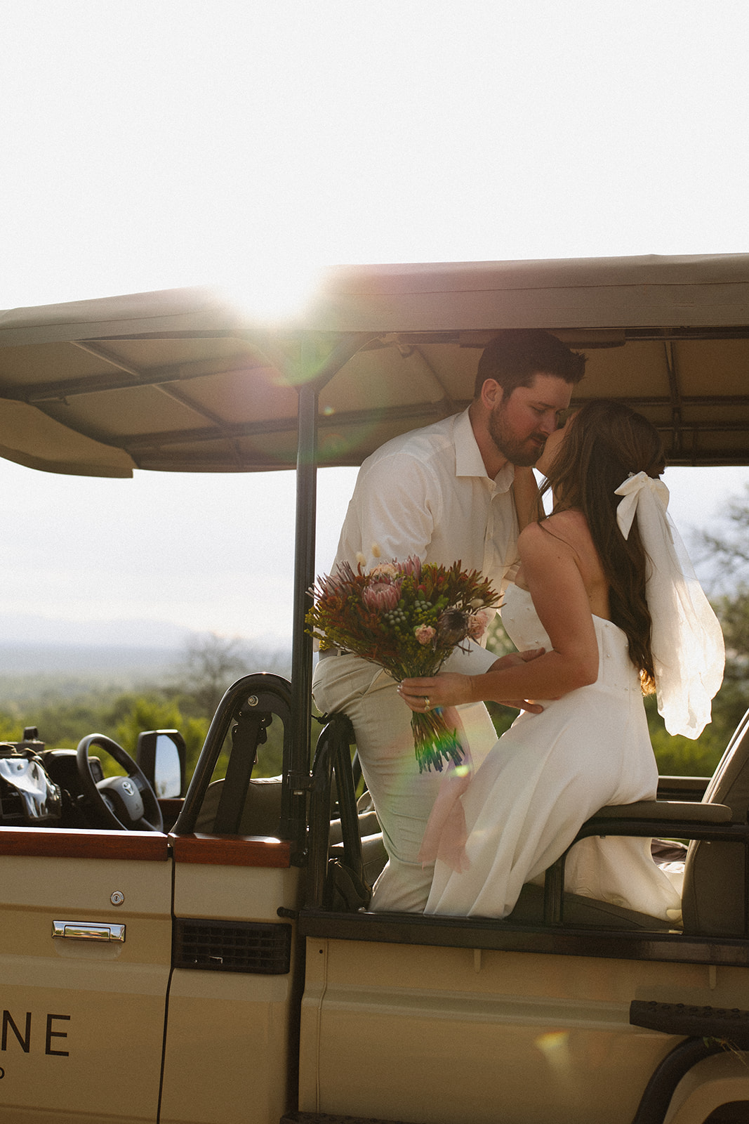 Bride and groom kissing on safari jeep with bouquet in hand and glowing sunset light during their wedding day

