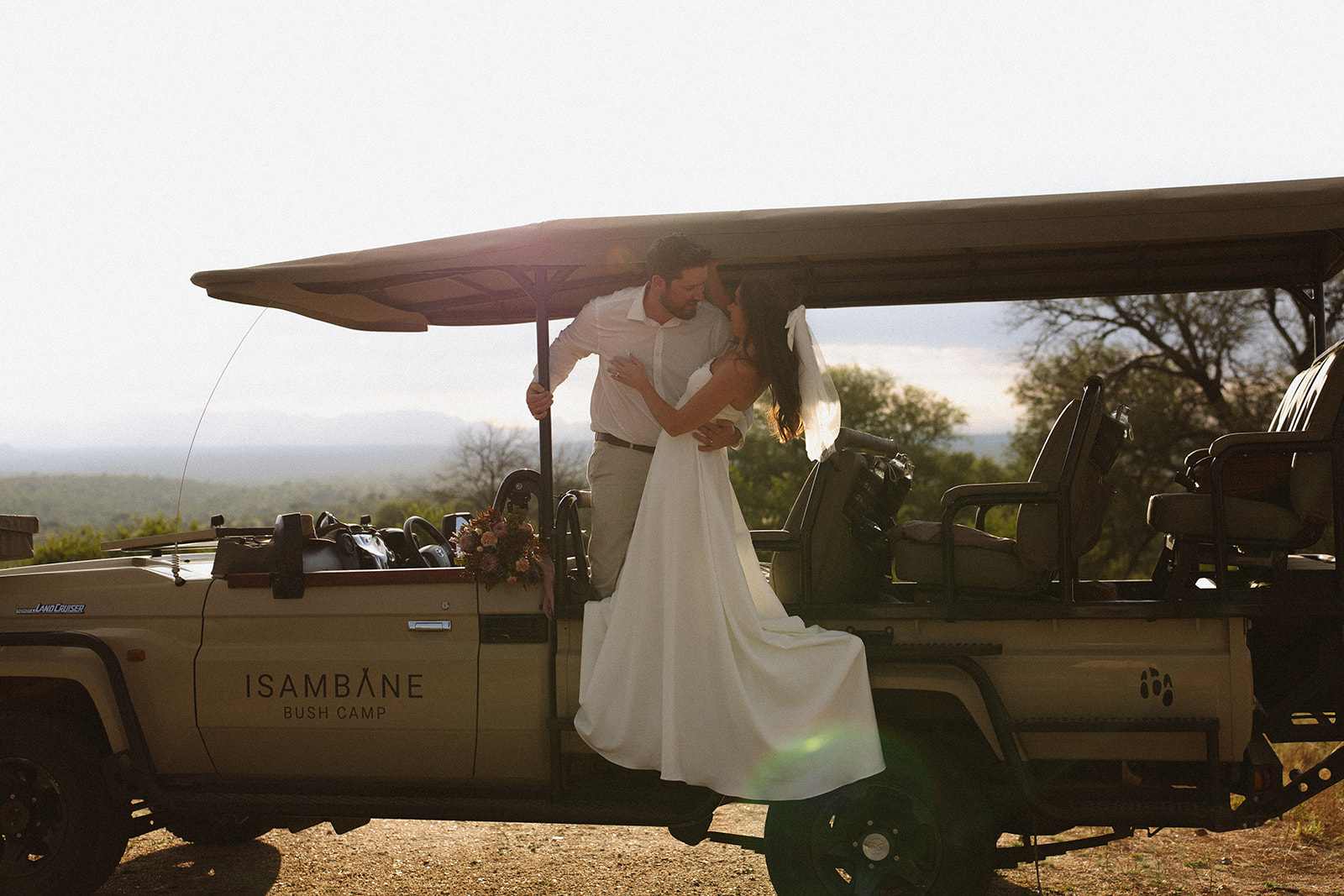 Bride and groom embracing on safari vehicle with bouquet and soft golden light during a romantic safari wedding packages moment
